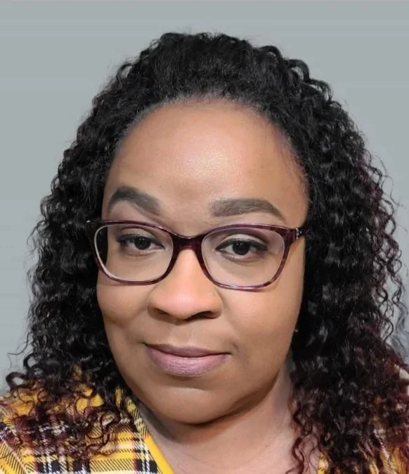 A woman with curly black hair wearing glasses and a yellow plaid top.