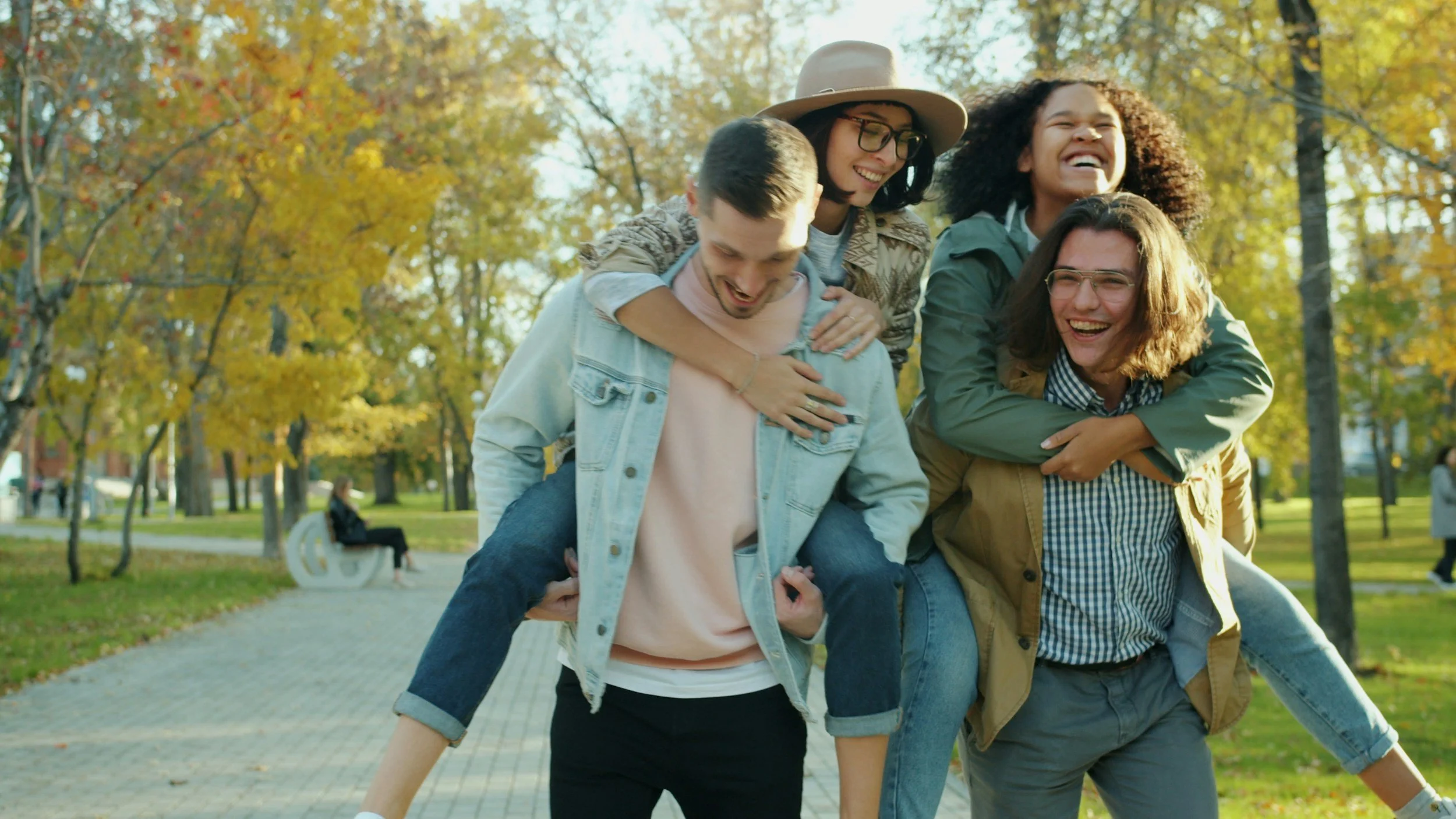 A group of five young adults enjoying happy marriages and a piggyback ride in a park with autumn trees in the background, smiling and laughing.