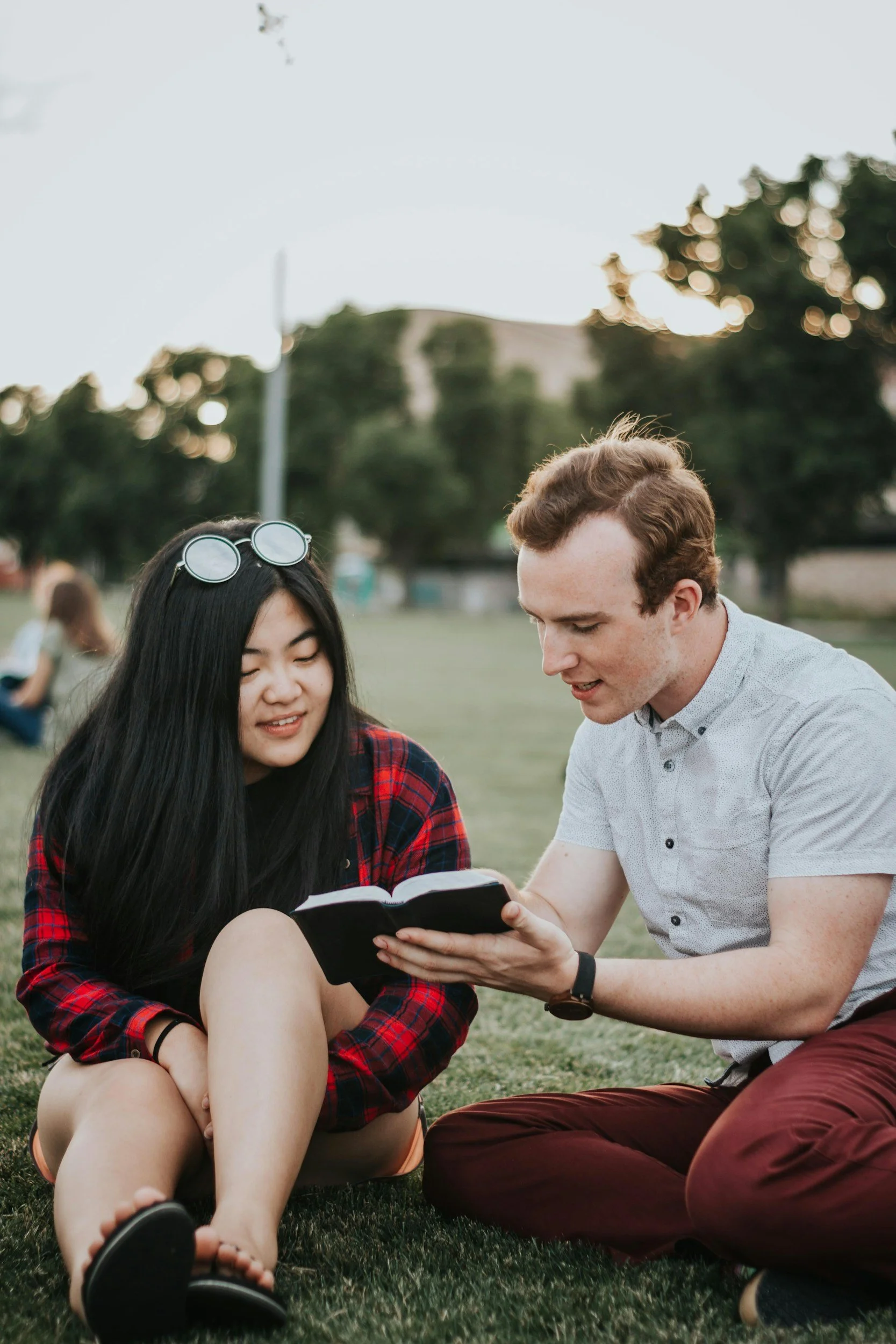 A young woman with long black hair, sunglasses on her head, and tan shorts sits on the grass with her legs crossed, looking at a book. A young man with red hair, wearing a light-colored short-sleeve shirt and brown pants, sits beside her holding the book together, both engaged in reading outside in a park with trees and a walkway in the background.