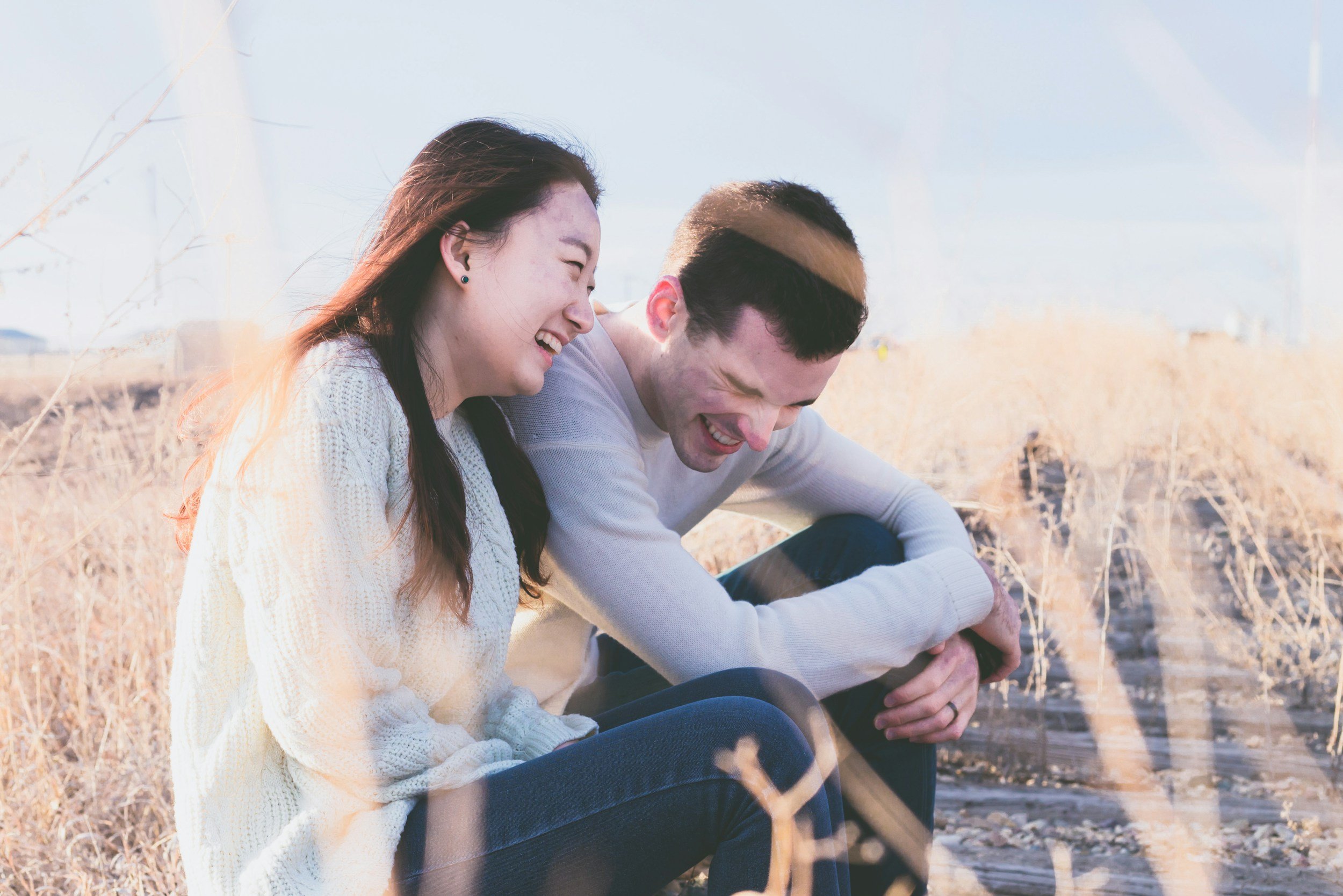 A young woman and a young man sitting together outdoors in a field, sharing a joyful moment and smiling while talking about their premarital counseling.