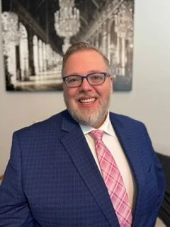 Portrait of a smiling man with glasses, beard, wearing a blue suit and pink tie, in front of black-and-white cityscape photograph.