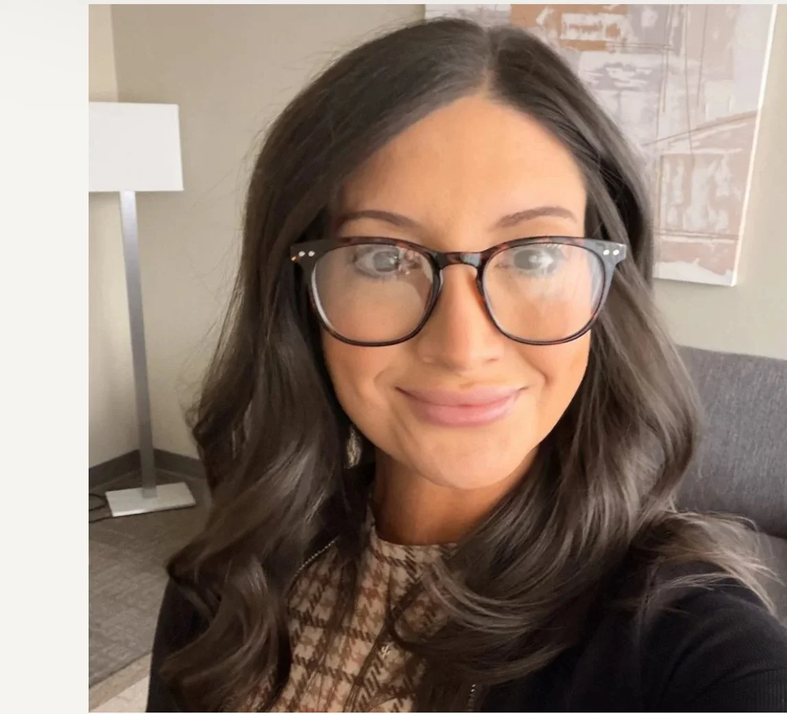 A woman with long wavy dark hair wearing glasses and a patterned top, sitting in a room with a gray sofa, a white wall with artwork, and a floor lamp in the background.