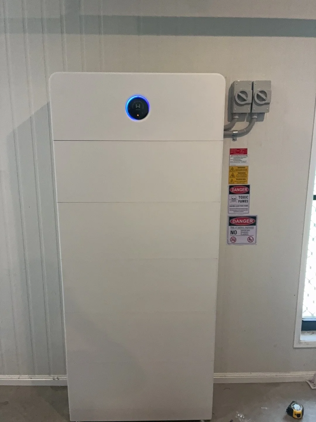 Large white electric vehicle charger station with a round digital display at the top, mounted on a wall next to electrical conduit and warning labels.