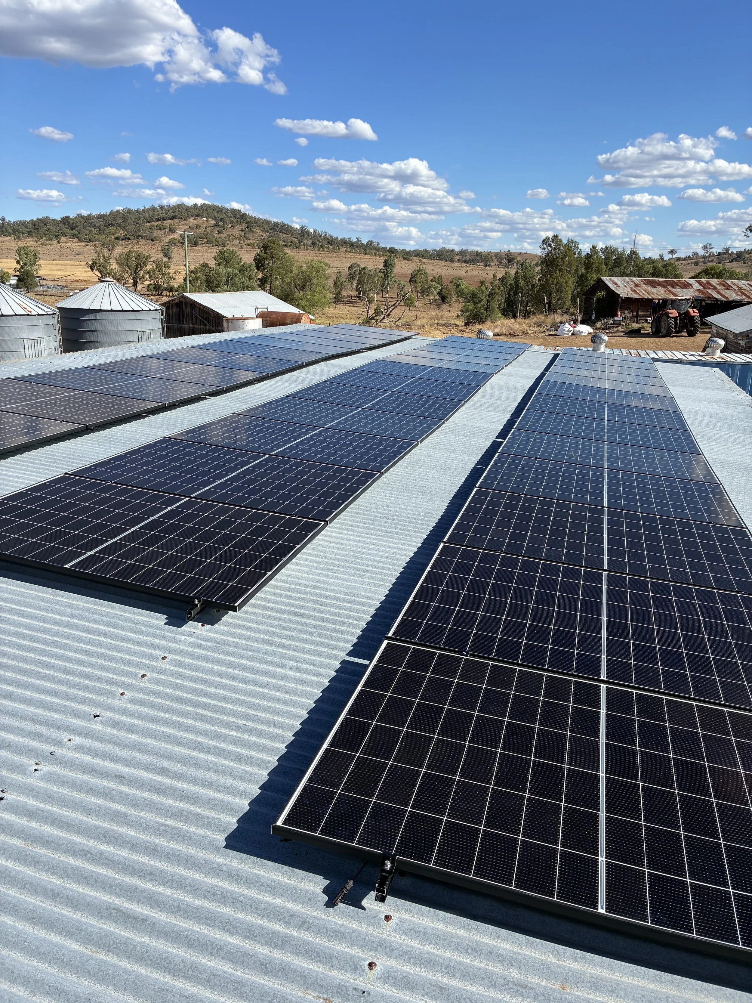 Solar panels installed on a metal roof with a farm landscape and blue sky with scattered clouds in the background.