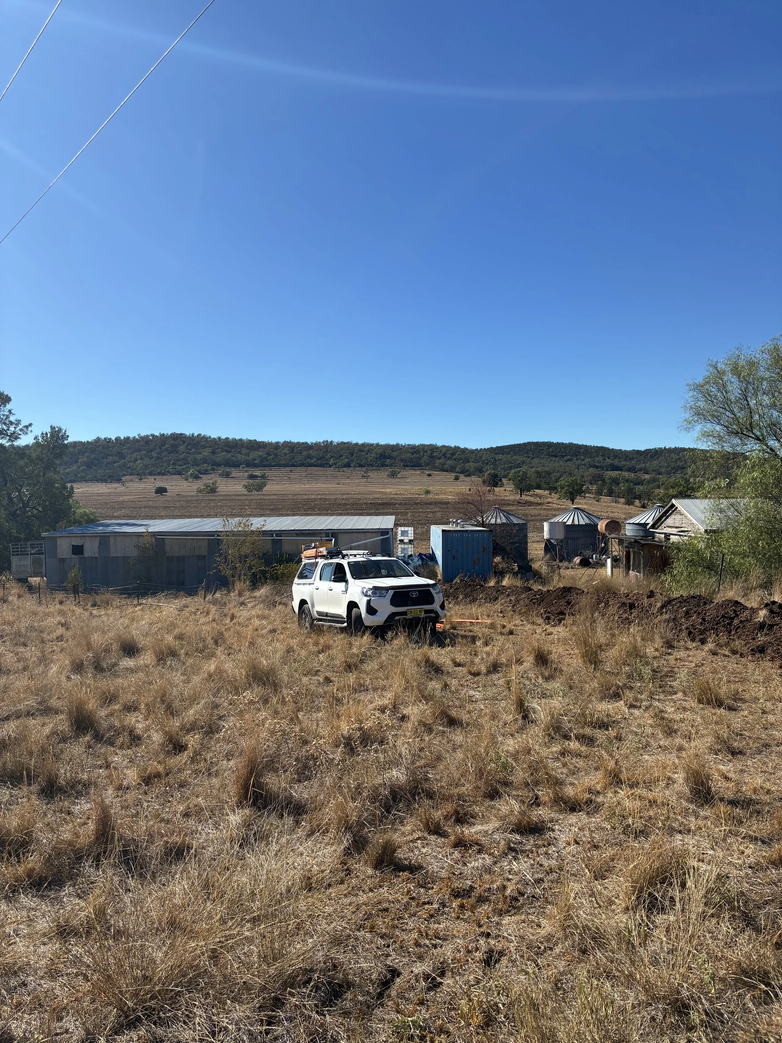 A rural scene with dry grass in the foreground, old metal buildings and silos, rolling hills with trees in the background, and a clear blue sky with a faint cloud streak.