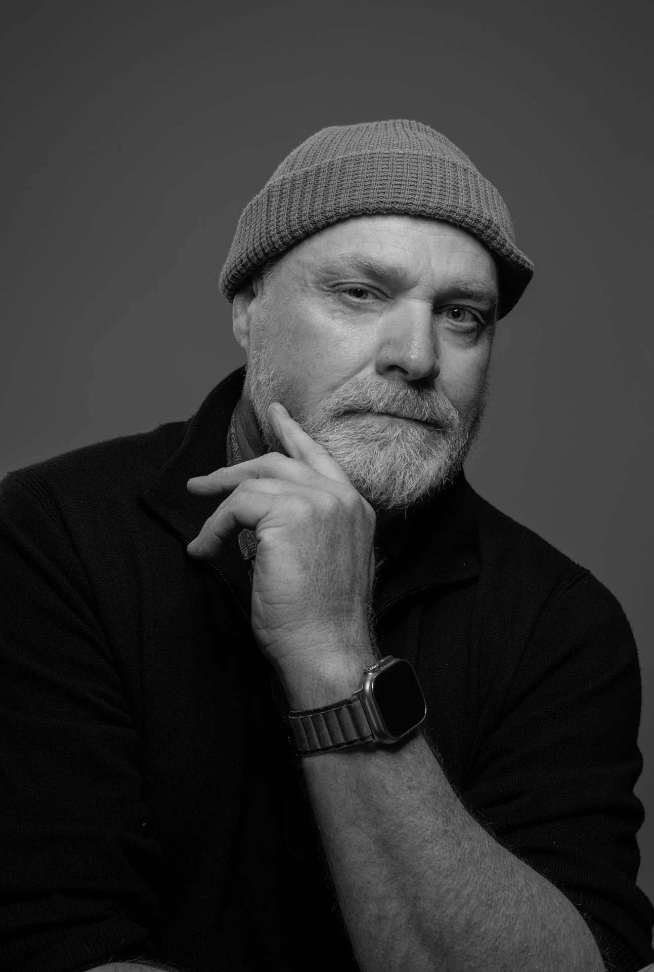 A black and white portrait of a middle-aged man with a beard, wearing a beanie and a smartwatch, looking at the camera with his hand resting on his chin.