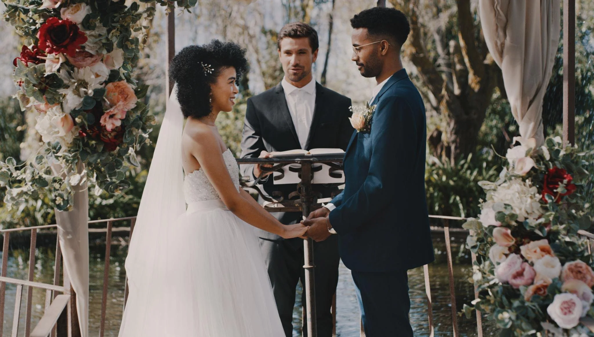 A couple getting married outdoors, holding hands and exchanging vows, under a floral arch with pink and white flowers, with an officiant standing behind them, in front of a body of water and trees.