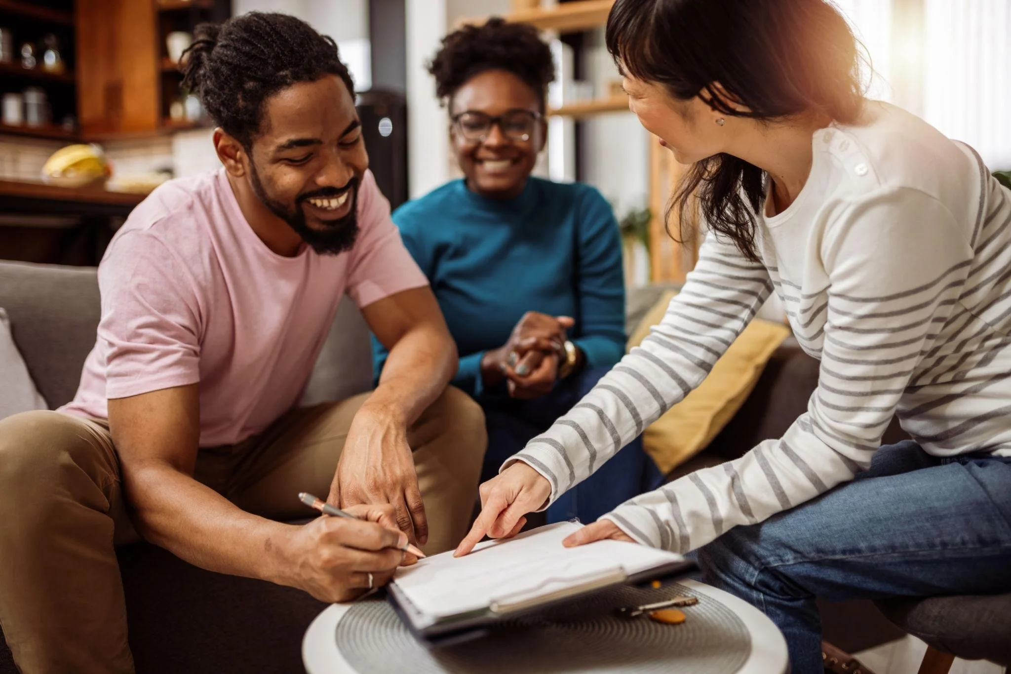 Three people are gathered around a table, with two women and one man, smiling and engaging in a collaborative activity, possibly signing a document or reviewing a document, in a cozy living room.