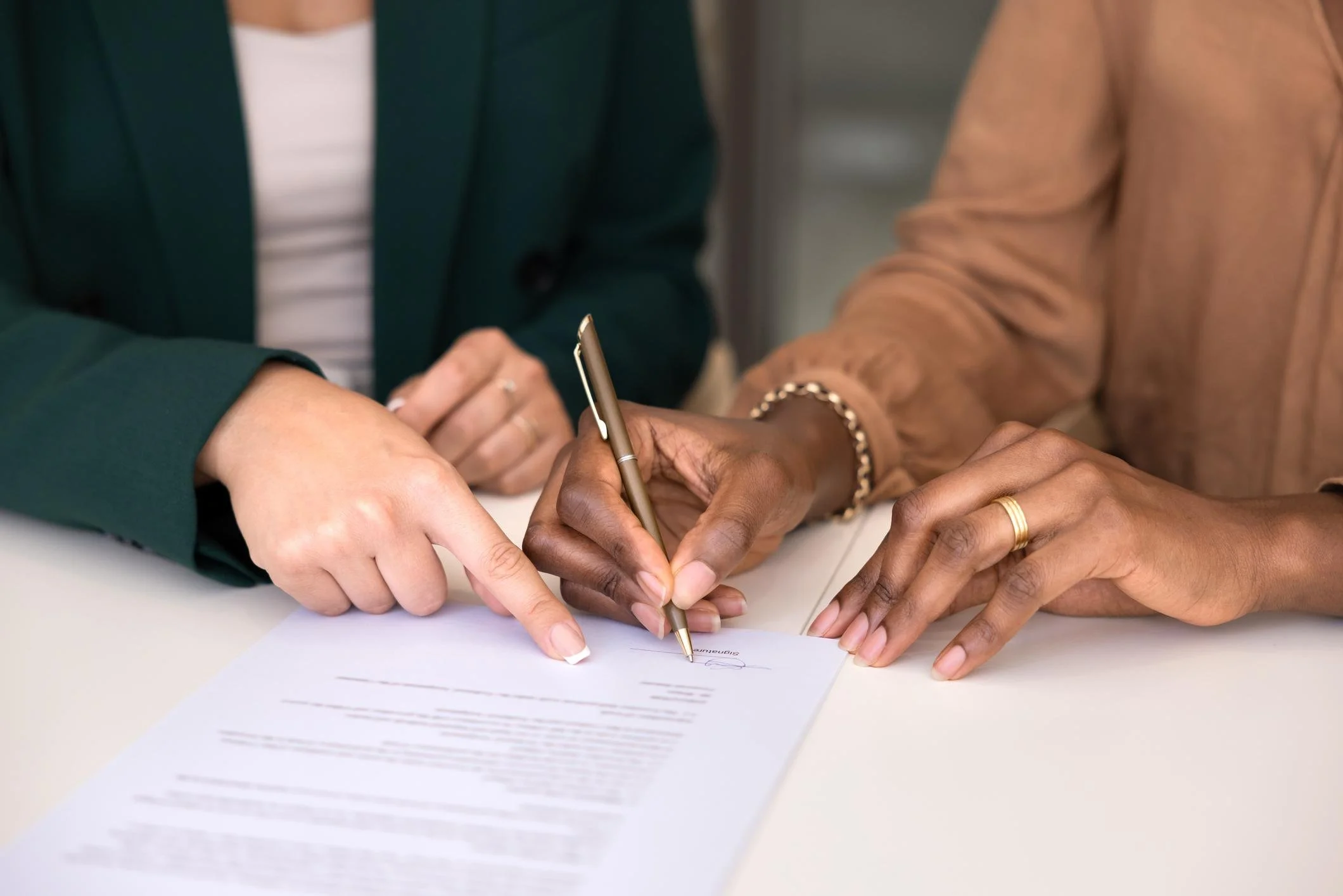 Two women are signing a document together, with one guiding and supporting the other's hand as they write.