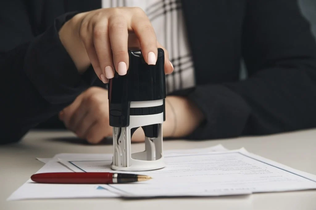 A person using a stamp to sign documents on a desk with a pen nearby.