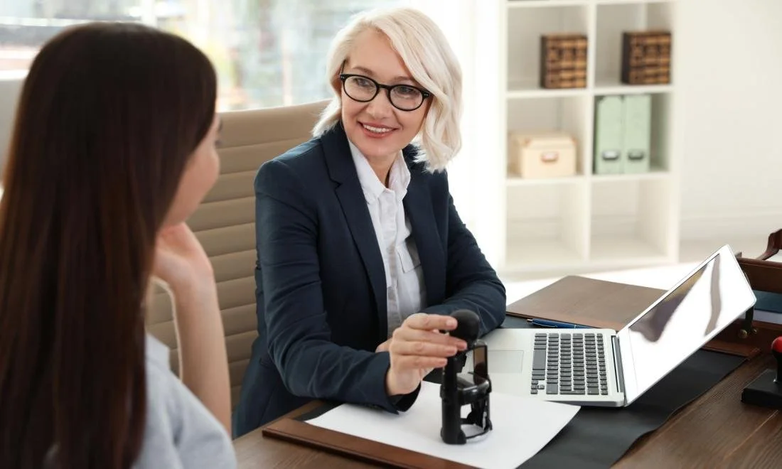 A professional woman with blonde hair and glasses talking to a woman with brown hair in an office setting. The woman with blonde hair is holding a stamp, with a laptop and office supplies on the desk.
