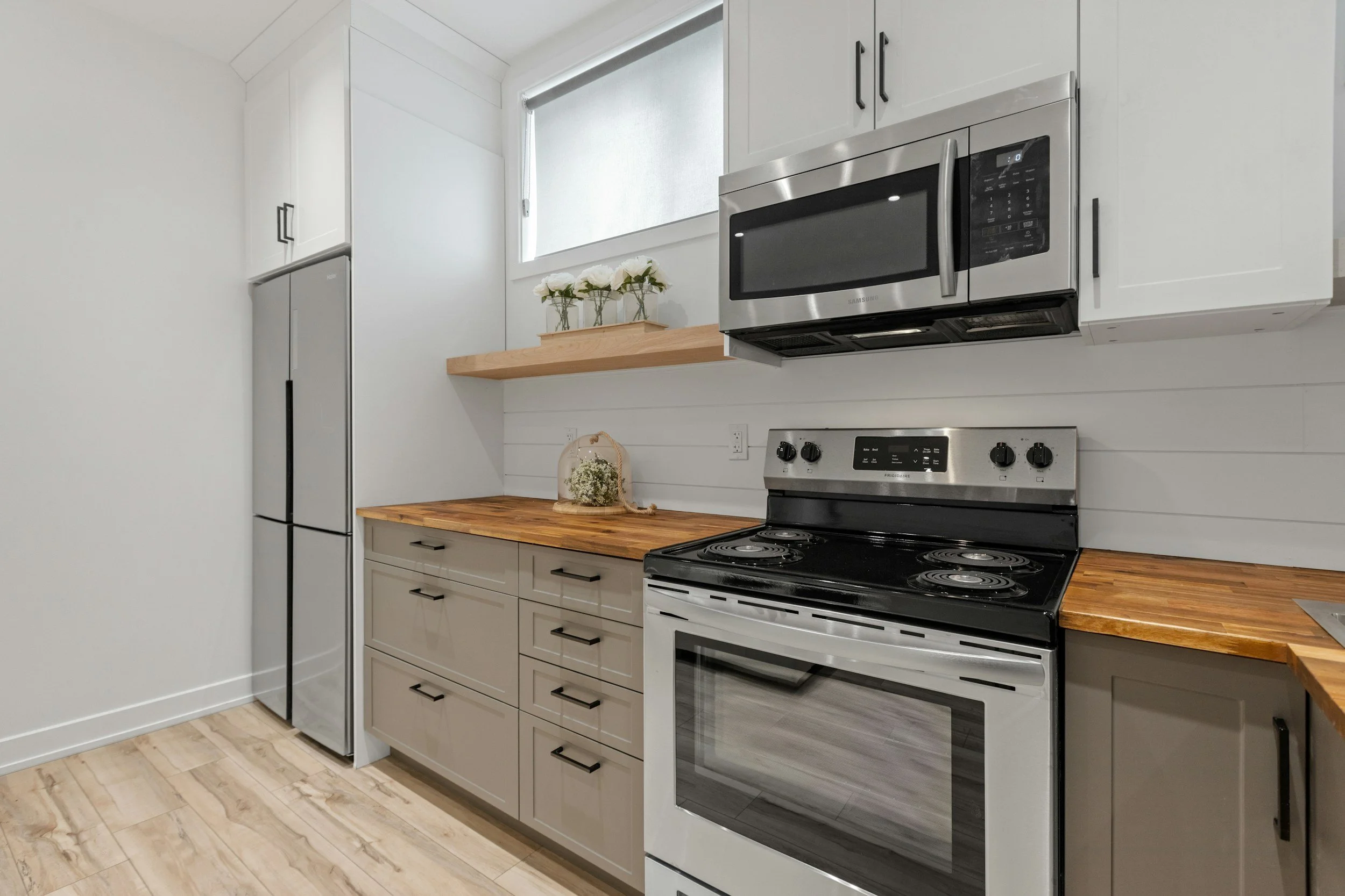A modern kitchen with white cabinets, a stainless steel stove with four burners, a stainless steel microwave, a tall refrigerator, a wooden countertop, and a window with a plant. The floor is made of light-colored wood.
