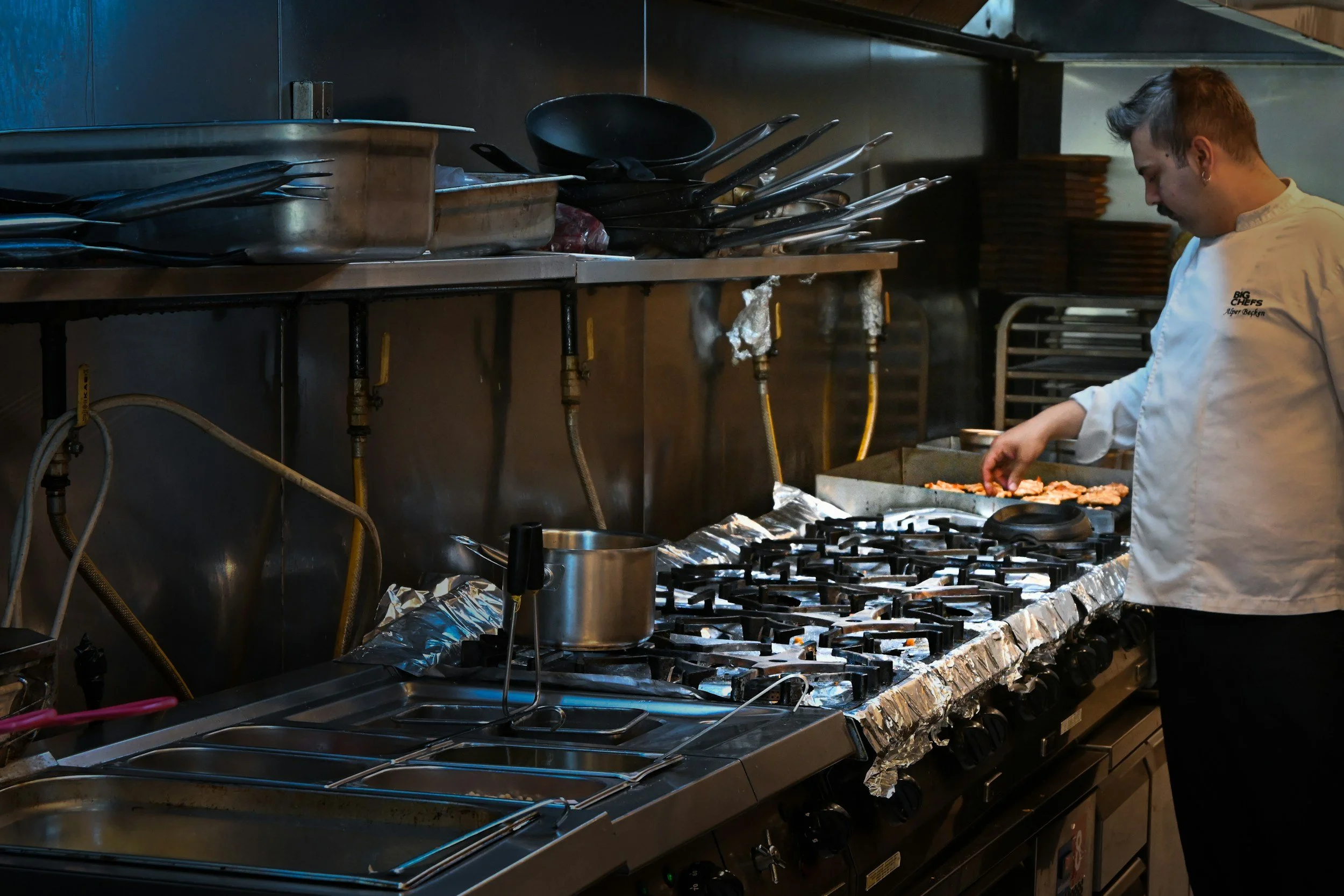 A chef in a white uniform cooking on large commercial stoves, with pans and utensils in a professional kitchen.