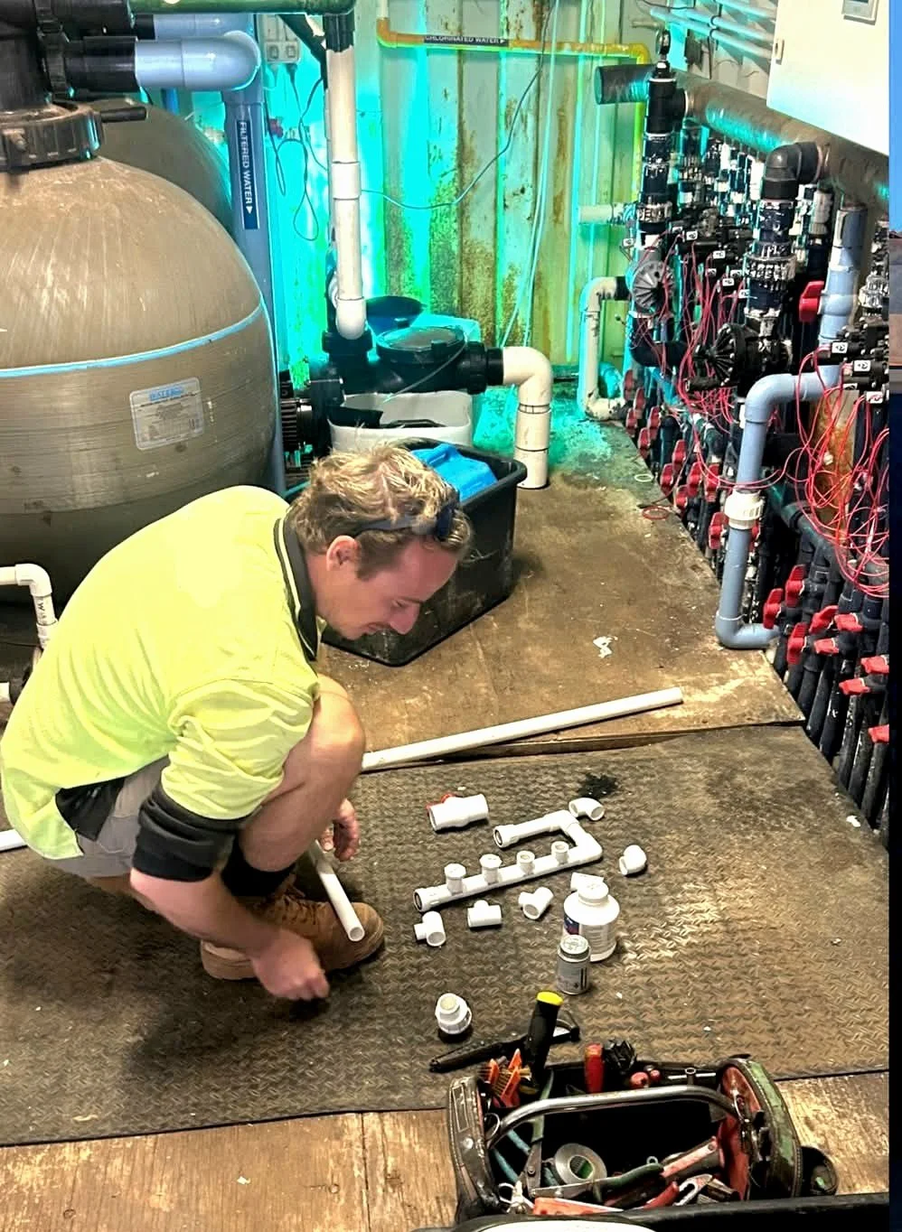 A man in a yellow shirt kneeling on a textured rubber mat, assembling white PVC pipes and fittings on the floor of a utility room with plumbing and a filtration system in the background.