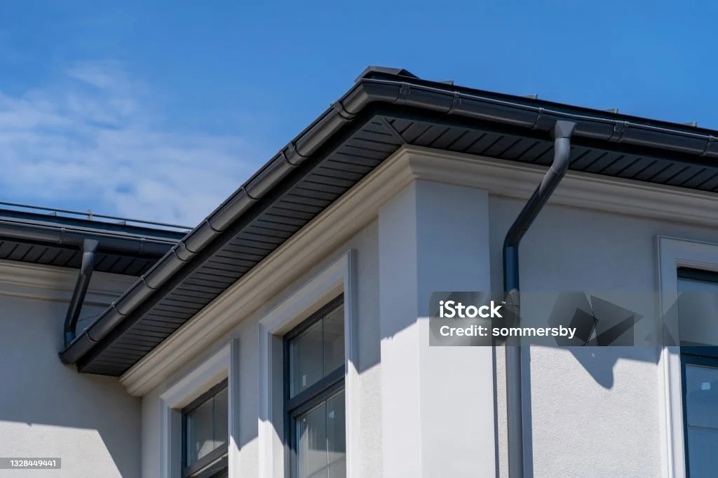 Close-up of a white house corner with a dark gray roof, gutters, and a rain downspout against a blue sky.