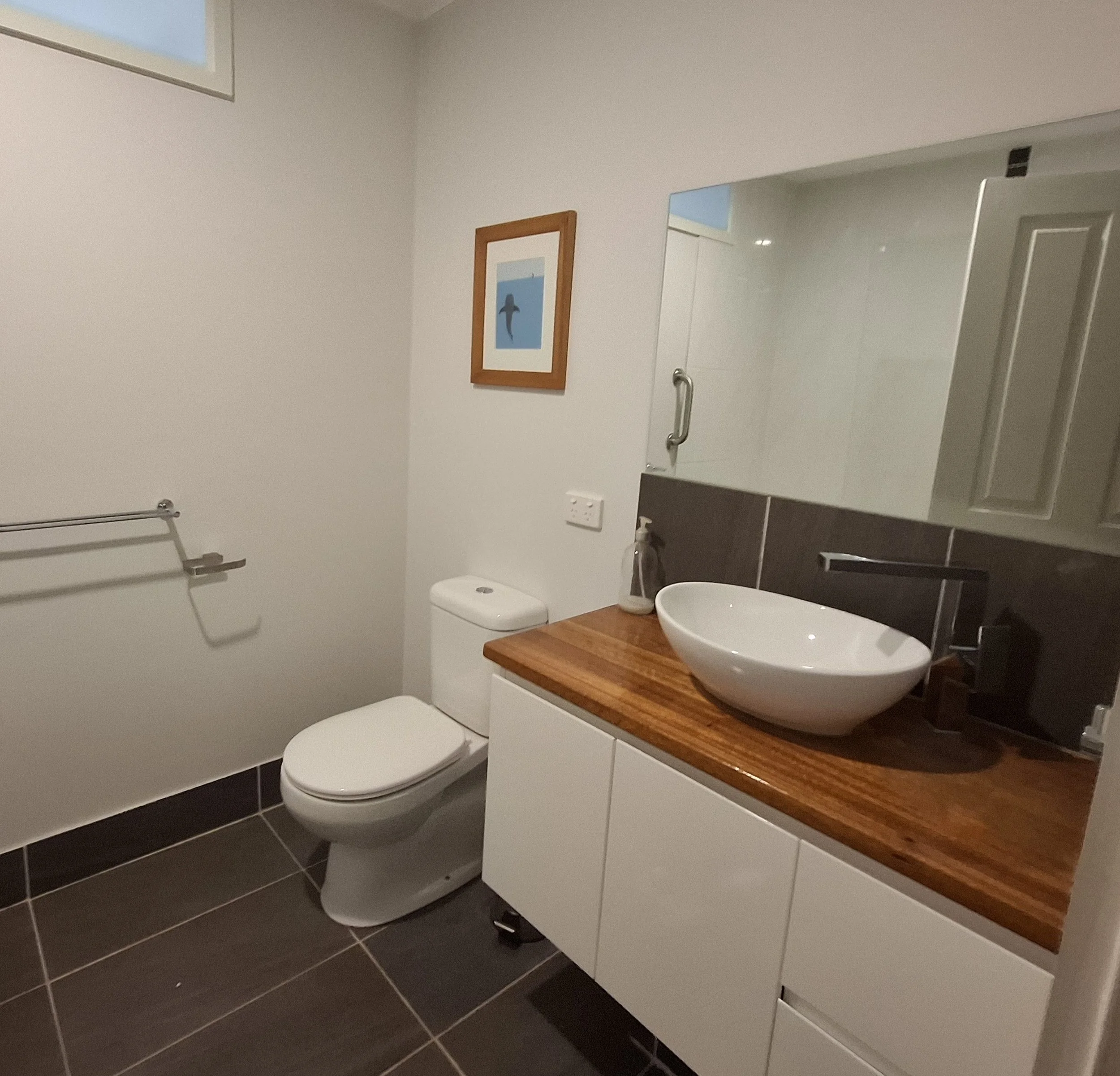 A modern bathroom with a white toilet, a white vessel sink on a wooden countertop, a black cabinet, and framed artwork of a dolphin on the white wall.