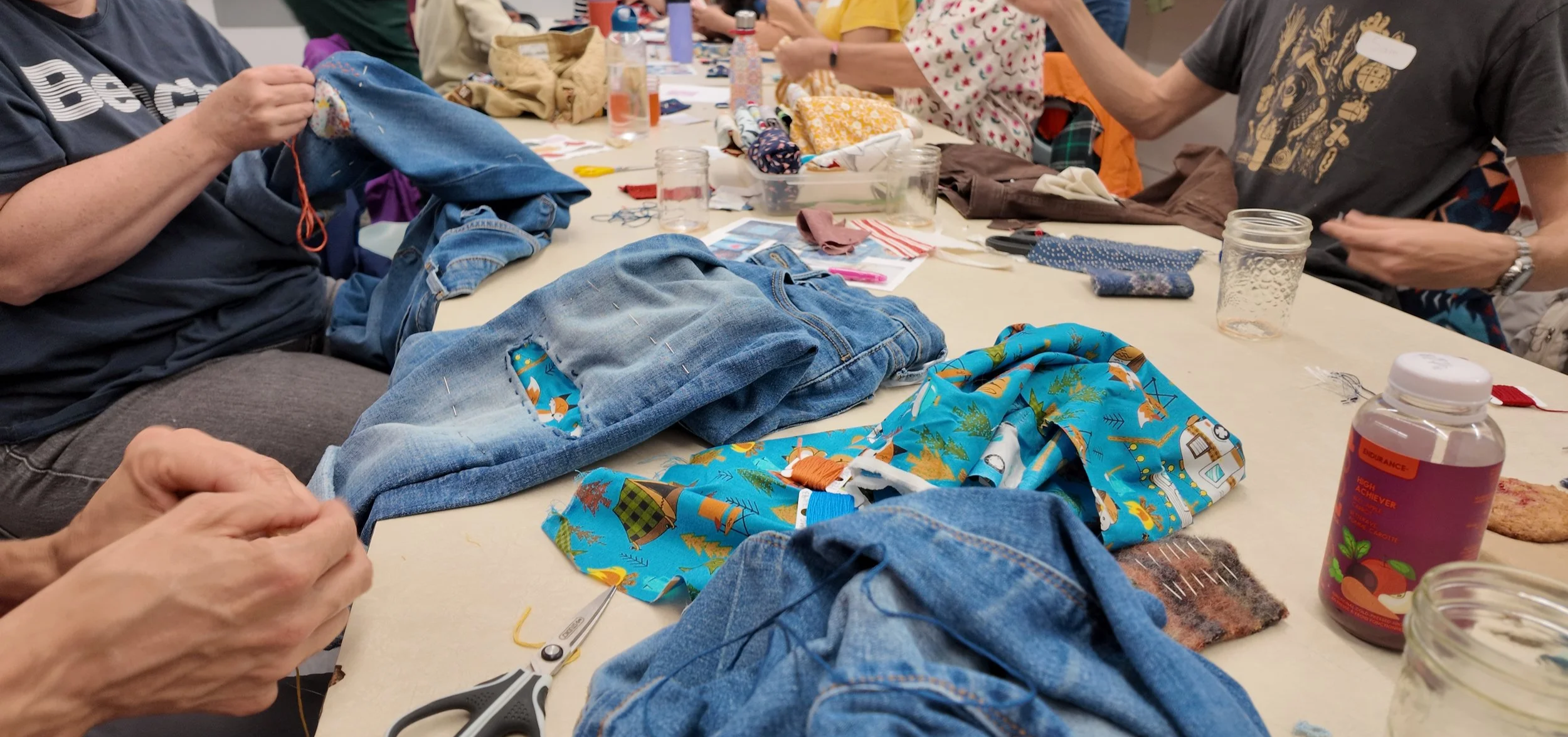People sitting at a table working on sewing projects with fabric, scissors, and sewing tools scattered on the table.