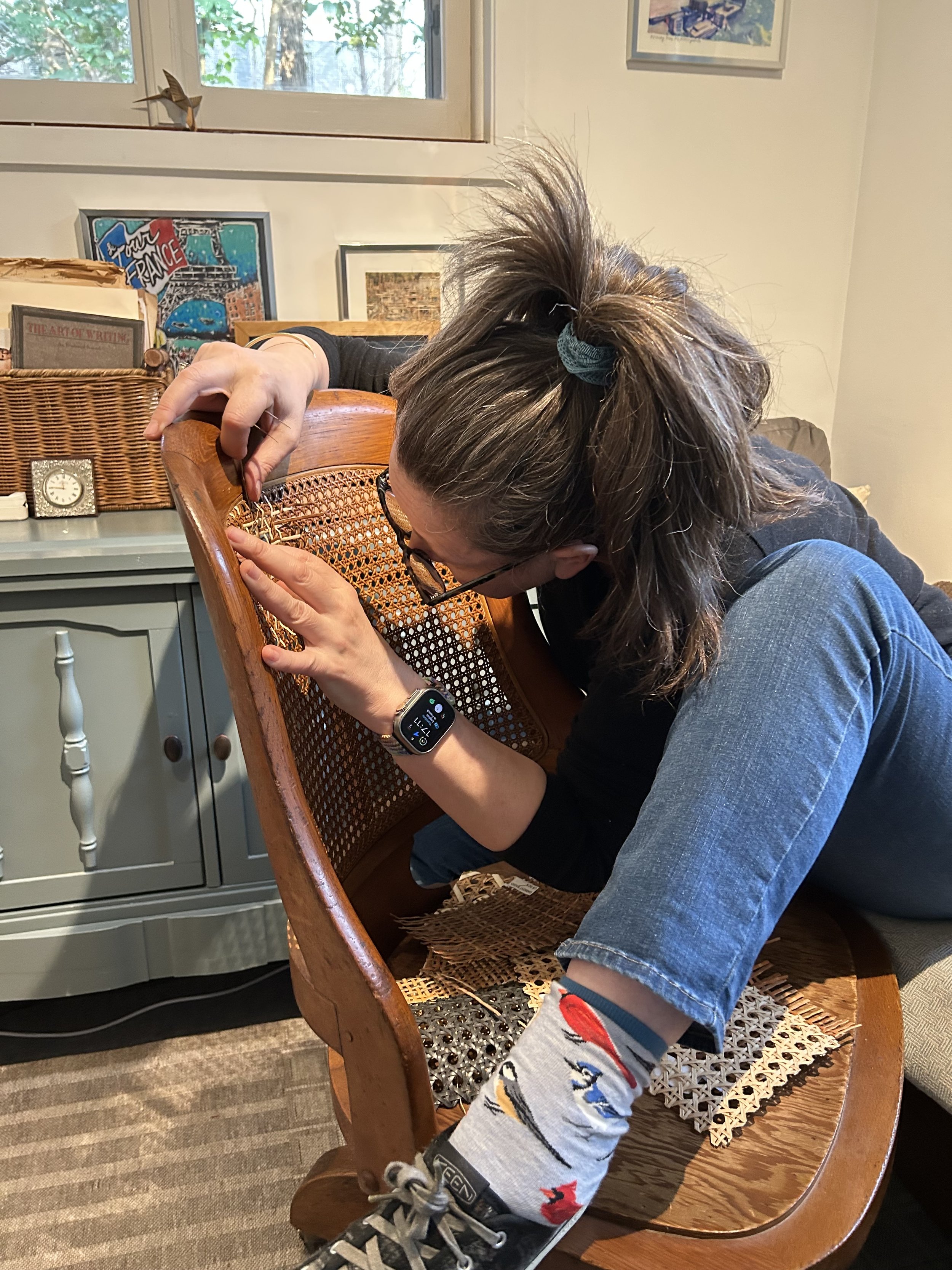 A woman with glasses and a ponytail is sitting on a wooden chair, leaning forward and fixing the caning on the back of the chair. Her foot is resting on the seat of the chair, showing a sock with colourful patterns.