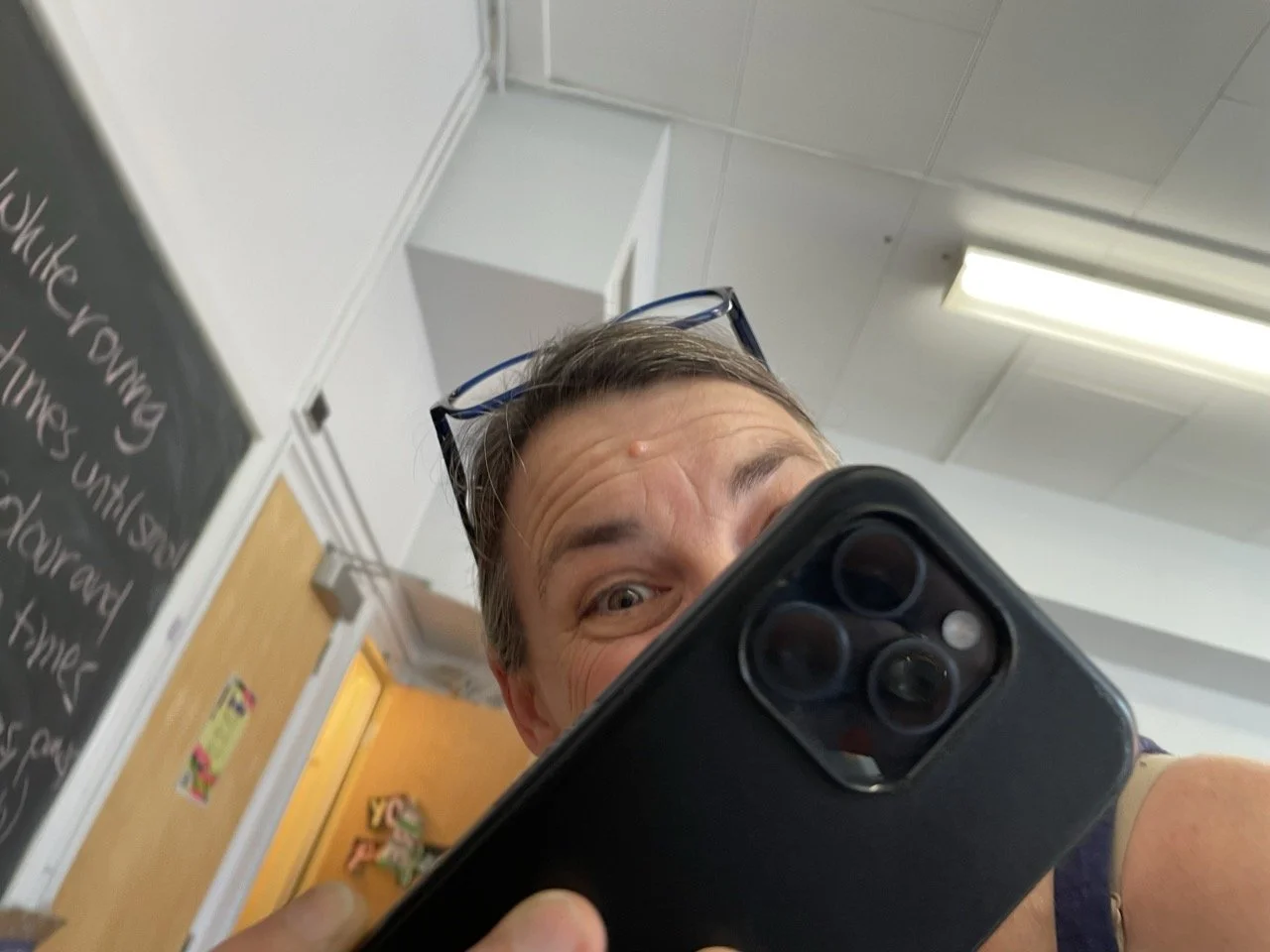 A woman in a classroom taking a picture with a smartphone. She has glasses on her head and he face is partially obscured, but you can tell that she is smiling.