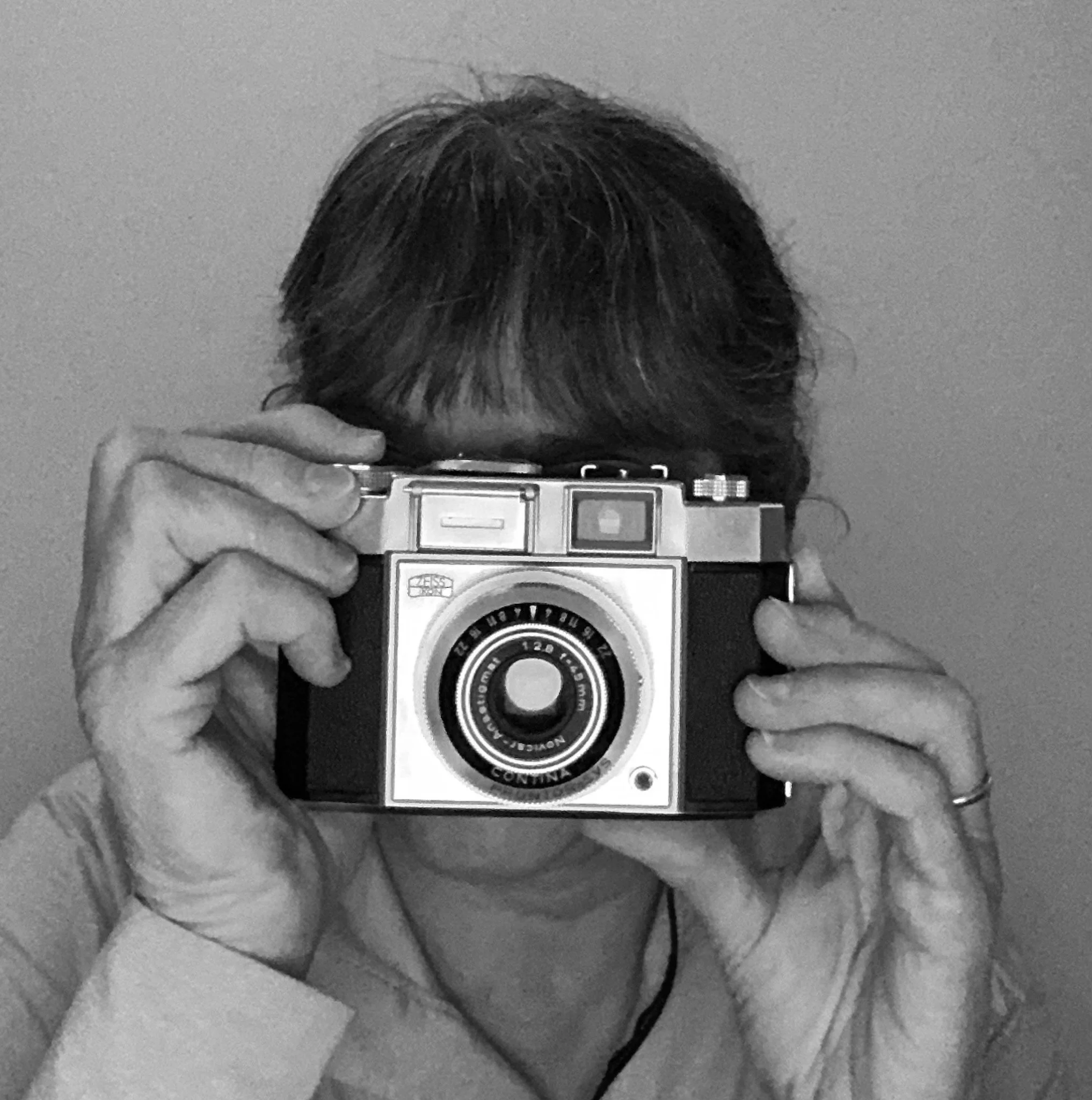 A woman with dark hair taking a selfie with a vintage camera in front of a mirror, partially obscuring her face.