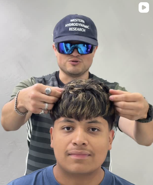 A man wearing sunglasses and a cap that says 'Western Hydrodynamic Research' is styling the hair of a young man in a blue shirt against a plain wall.