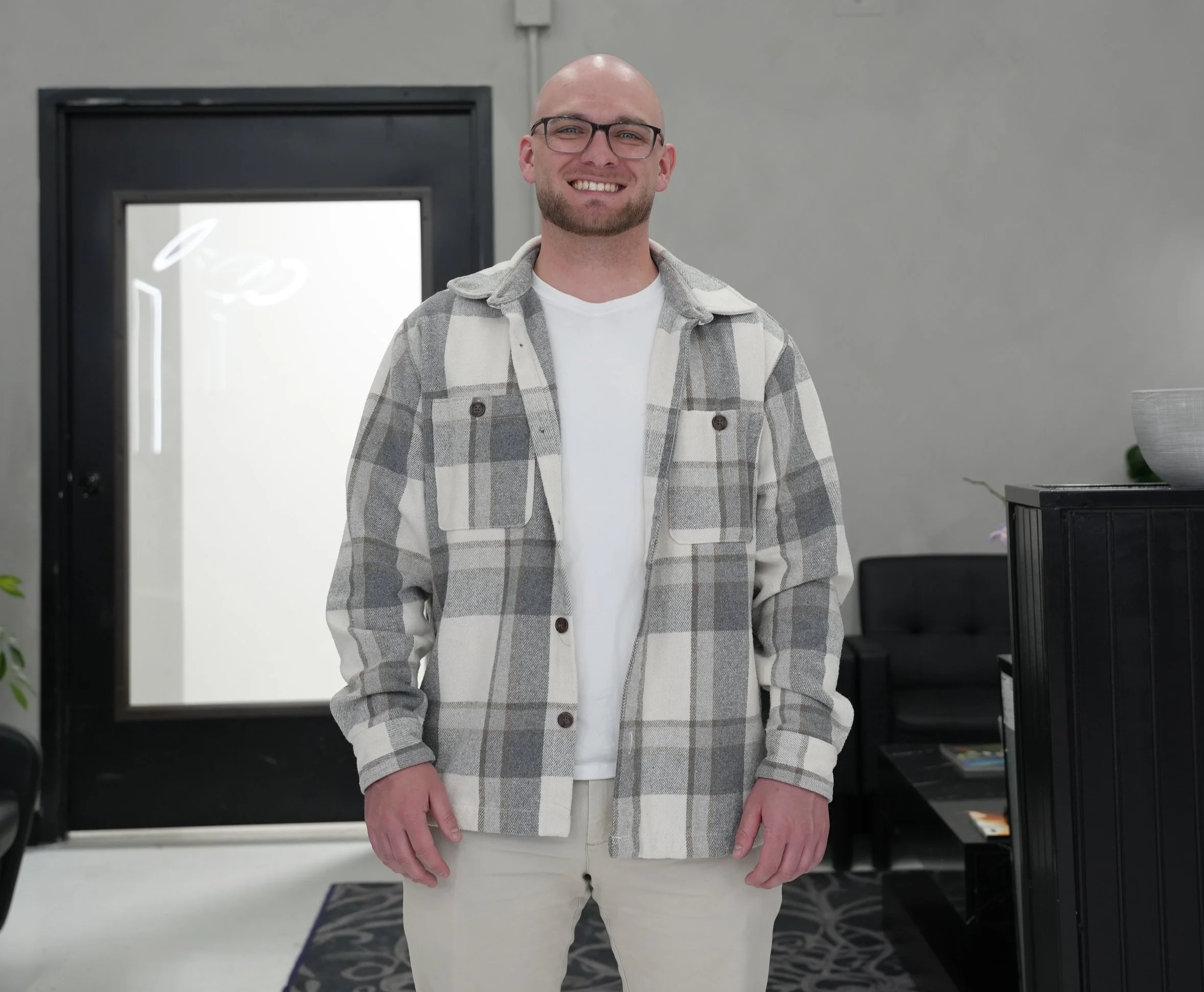 A smiling man with glasses, a beard, wearing a gray and white checkered jacket over a white shirt, standing in front of a gray wall and door with a glass window.