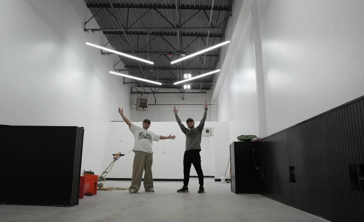 Two men standing inside a large, empty room with high ceilings. They are posing with their arms raised, one with two fingers up and the other with arms wide open. The room has industrial-style ceiling lights and some cleaning equipment on the floor.