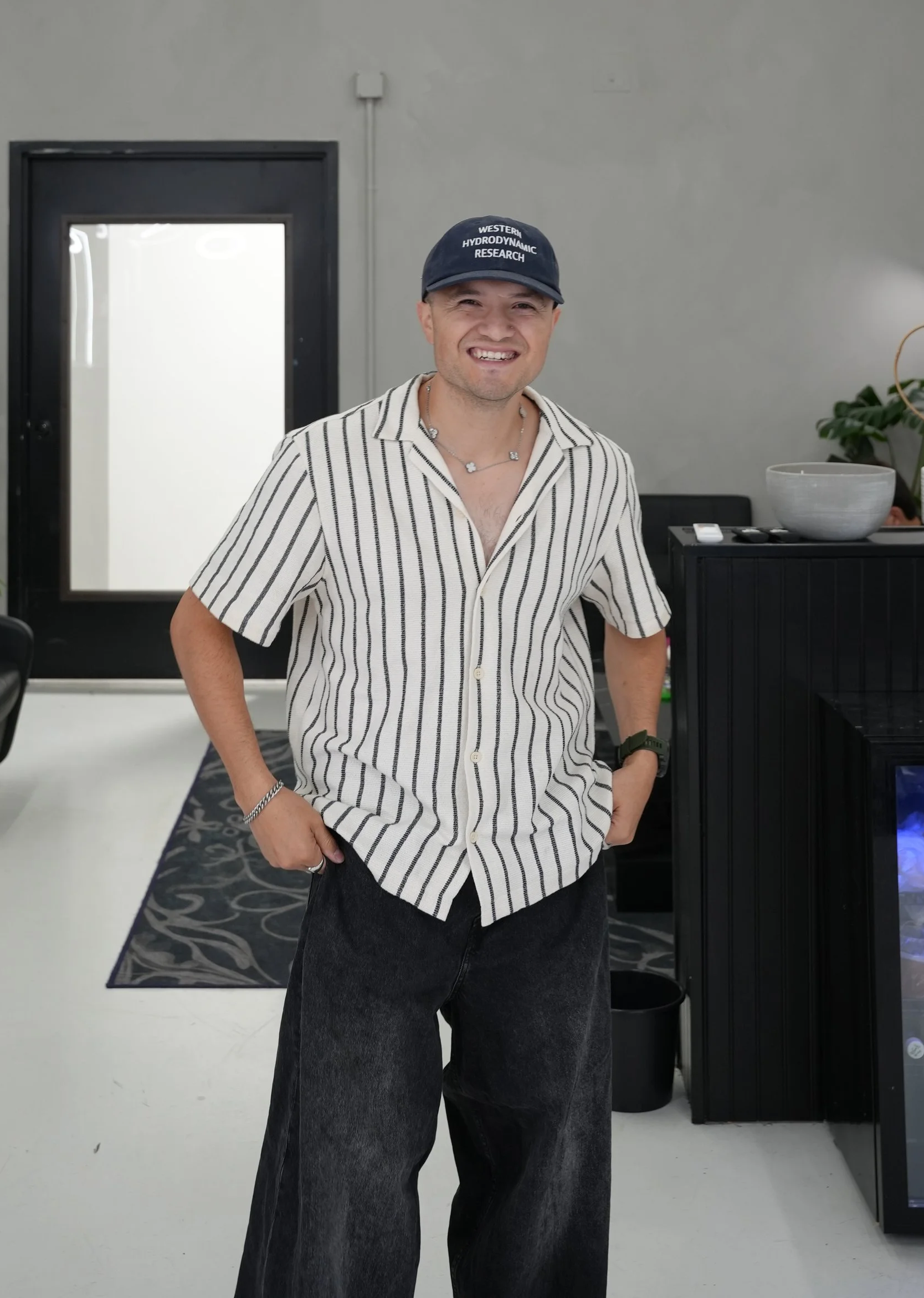 A smiling man in a striped shirt and black pants stands indoors, wearing a black cap that reads "Western Hydrodynamic Research," with his hands in his pockets.