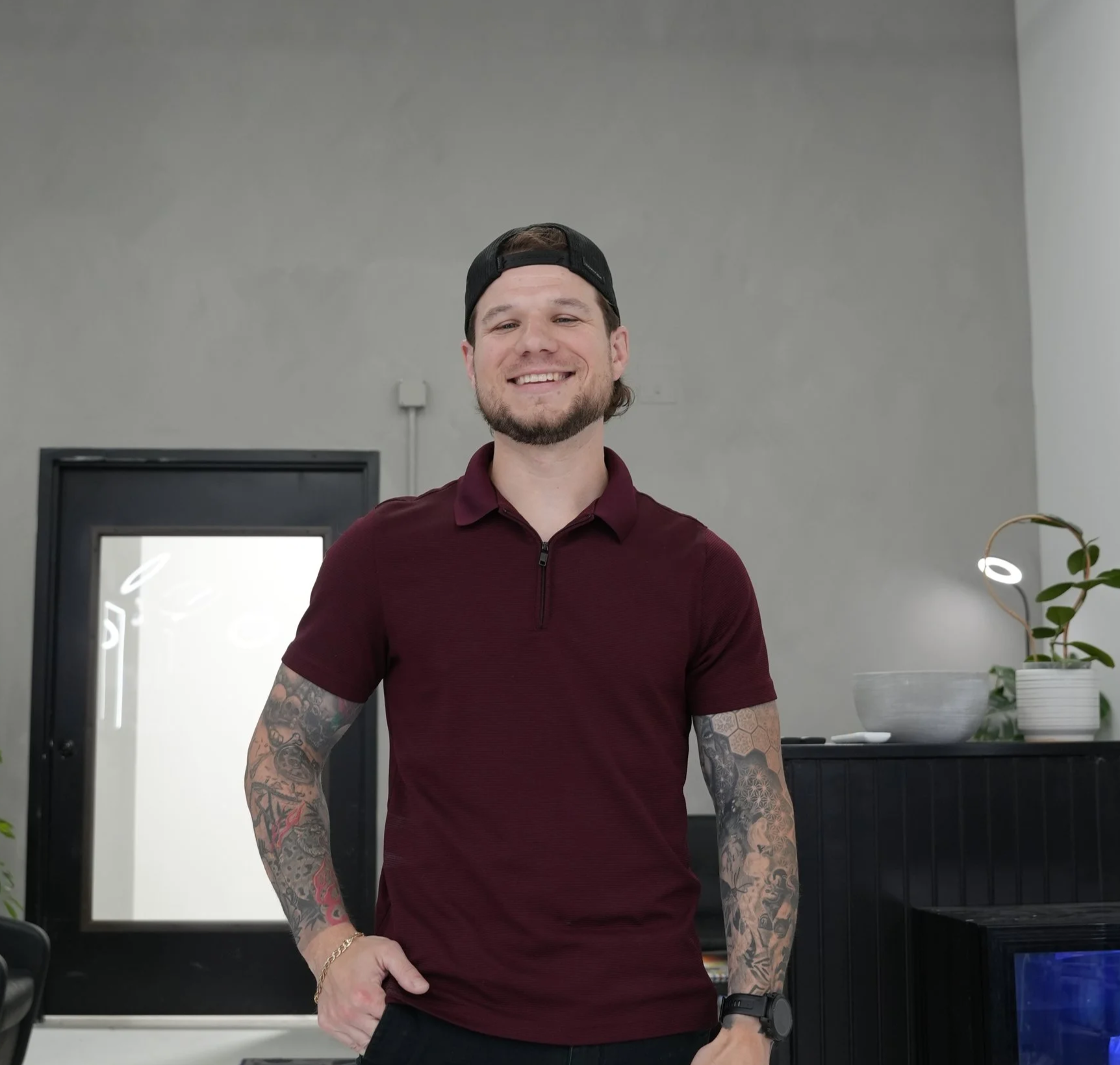 Smiling man with tattoos on arms, wearing a black cap backwards and a maroon polo shirt, standing indoors near a desk with a plant and a screen behind him.