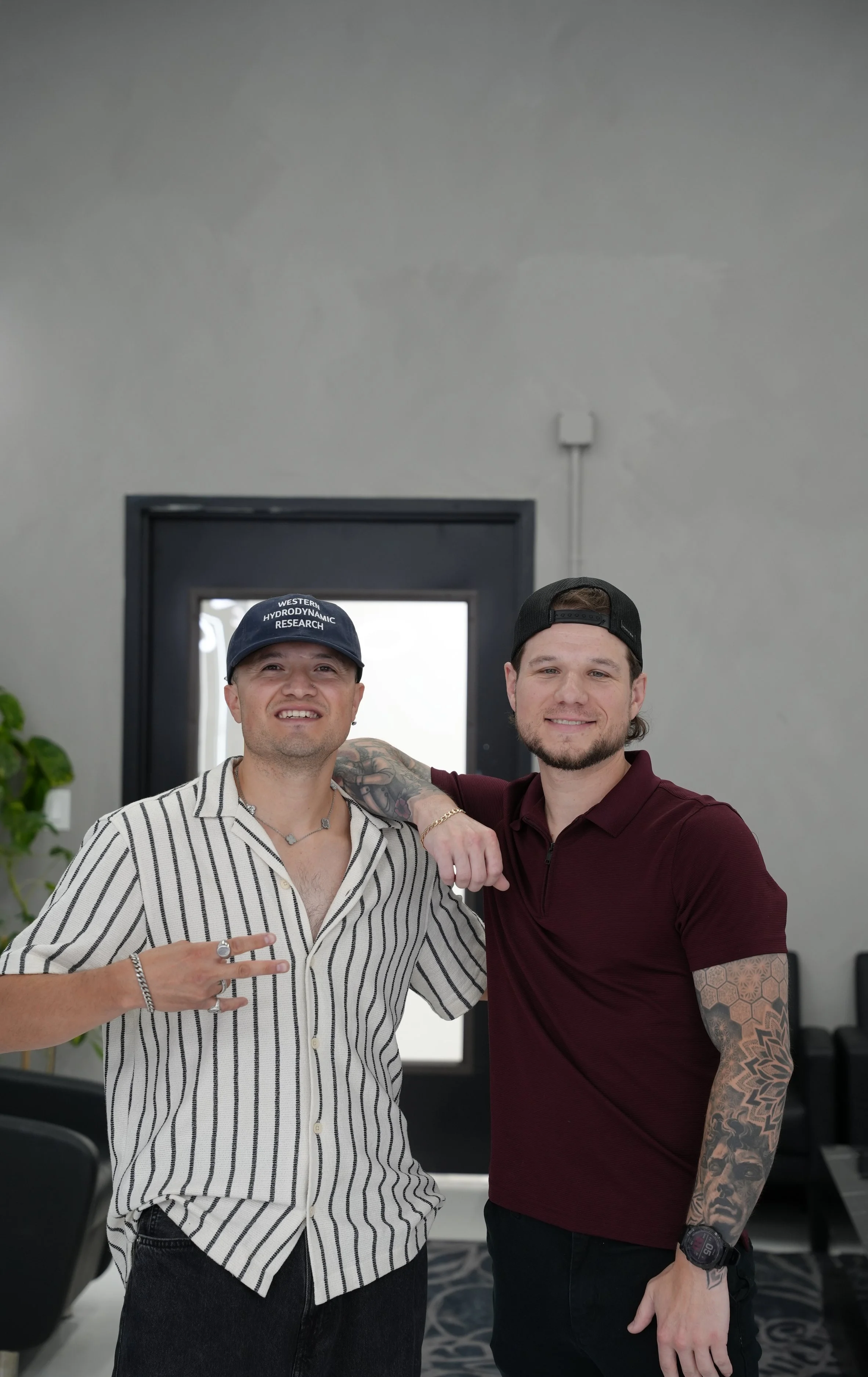 Two young men standing close together indoors, smiling at the camera. One is wearing a striped shirt, a cap that says 'Western Hydrodynamic Research,' and making a peace sign with his right hand. The other is dressed in a dark red polo shirt, a black cap worn backwards, has a tattooed sleeve on his left arm, and is smiling.