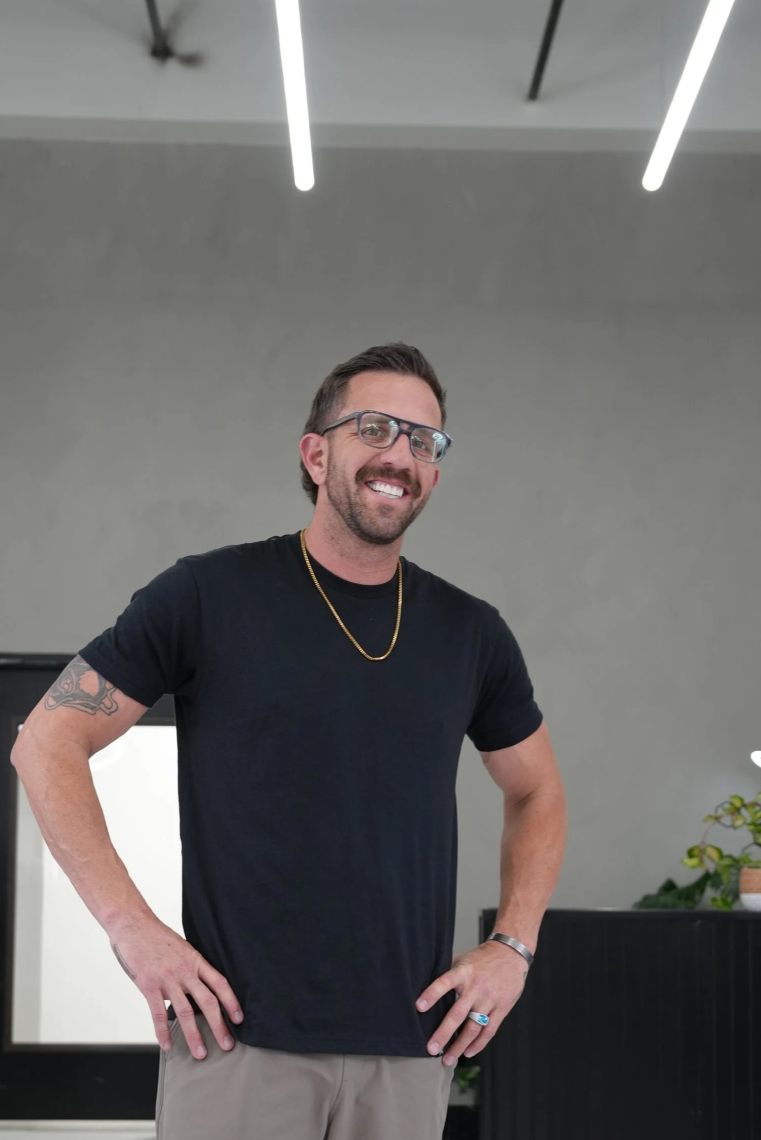 A smiling man with glasses, a black t-shirt, and beige pants standing with hands on hips in a modern interior space.