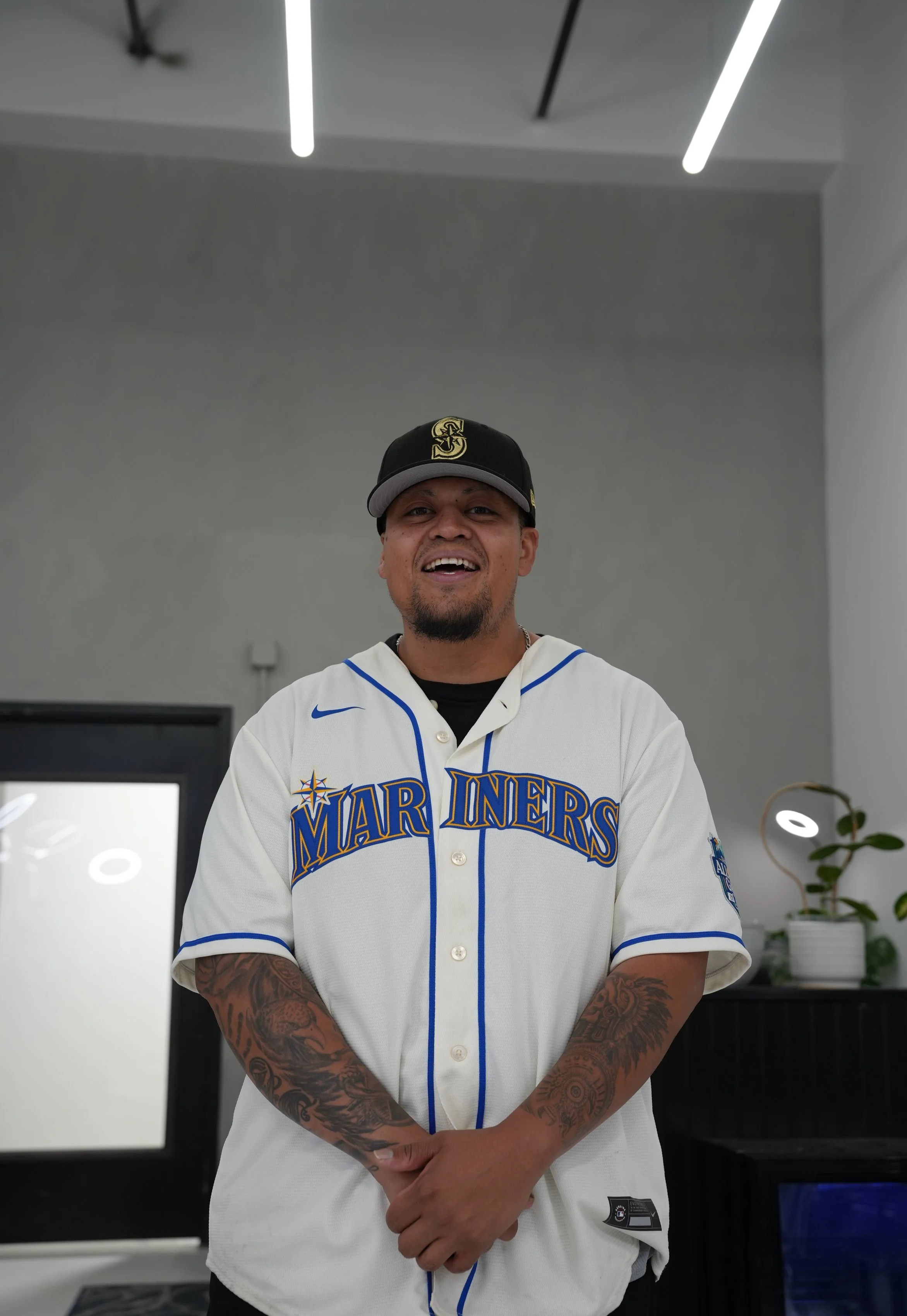 A man is smiling and wearing a Marin Mariners baseball jersey and a Seattle Mariners cap, standing indoors with a plain gray wall and minimal decor in the background.