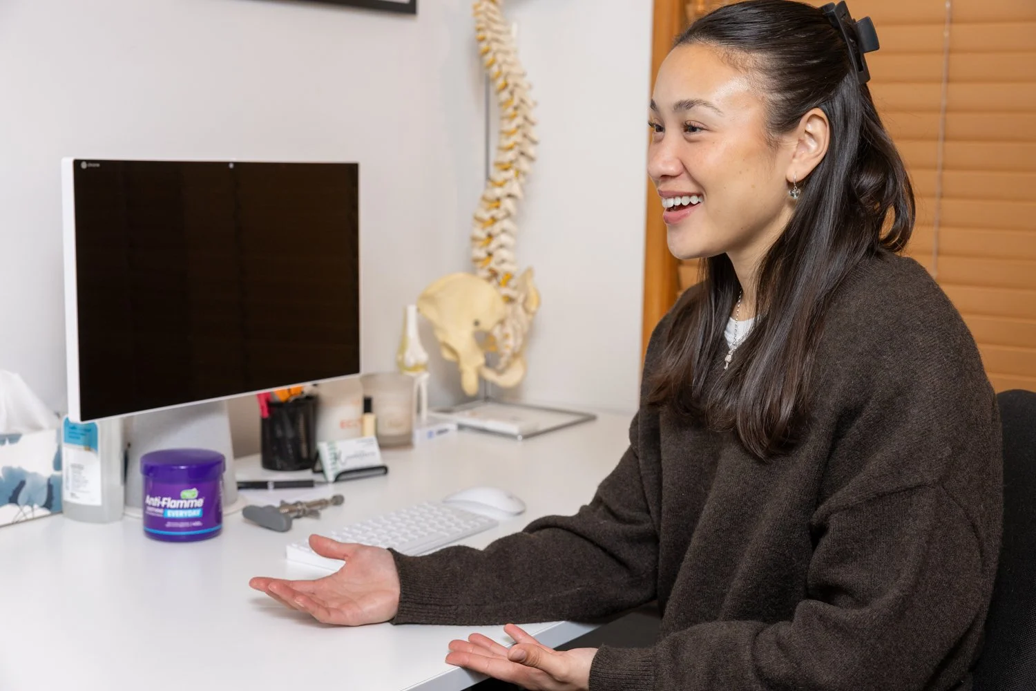 A woman with long dark hair, wearing a dark sweater and hair clip, sitting at a desk in a chiropractic office. The desk has a computer and various chiropractic equipment, including a model of the human spine in the background.