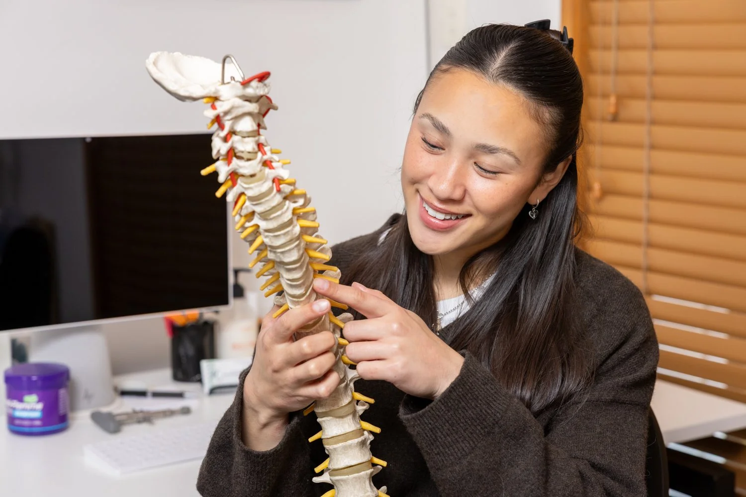 Woman smiling and pointing at a model of a human spine with yellow nerves in an office setting.