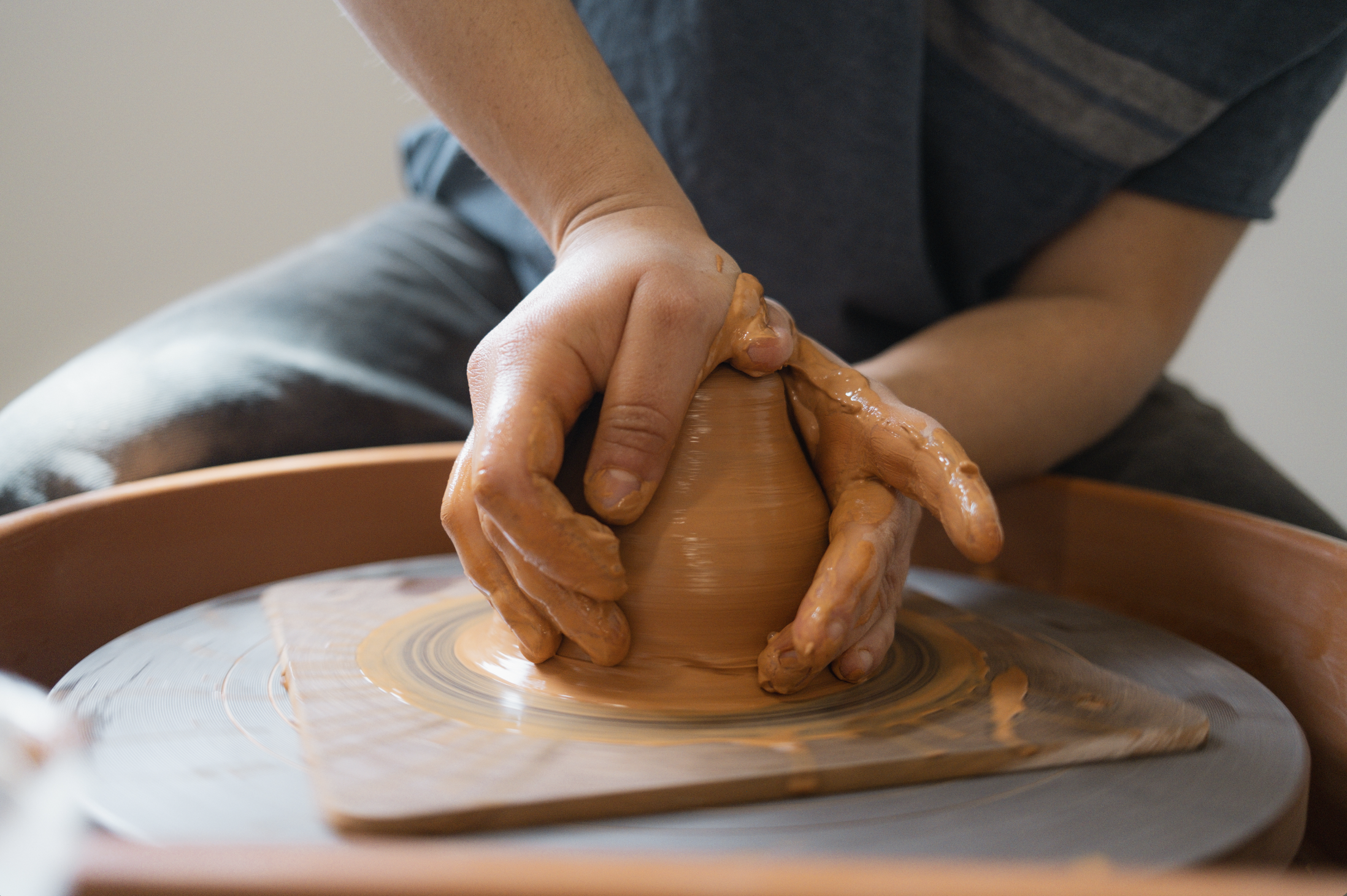 A person shaping a clay pot on a pottery wheel, with clay on their hands.
