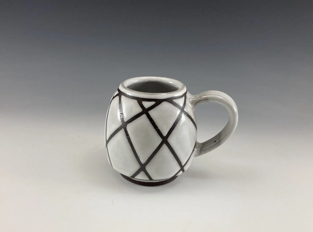 A small ceramic mug with a white background and black geometric lines forming diamond shapes, with a rounded handle and a narrow opening at the top.