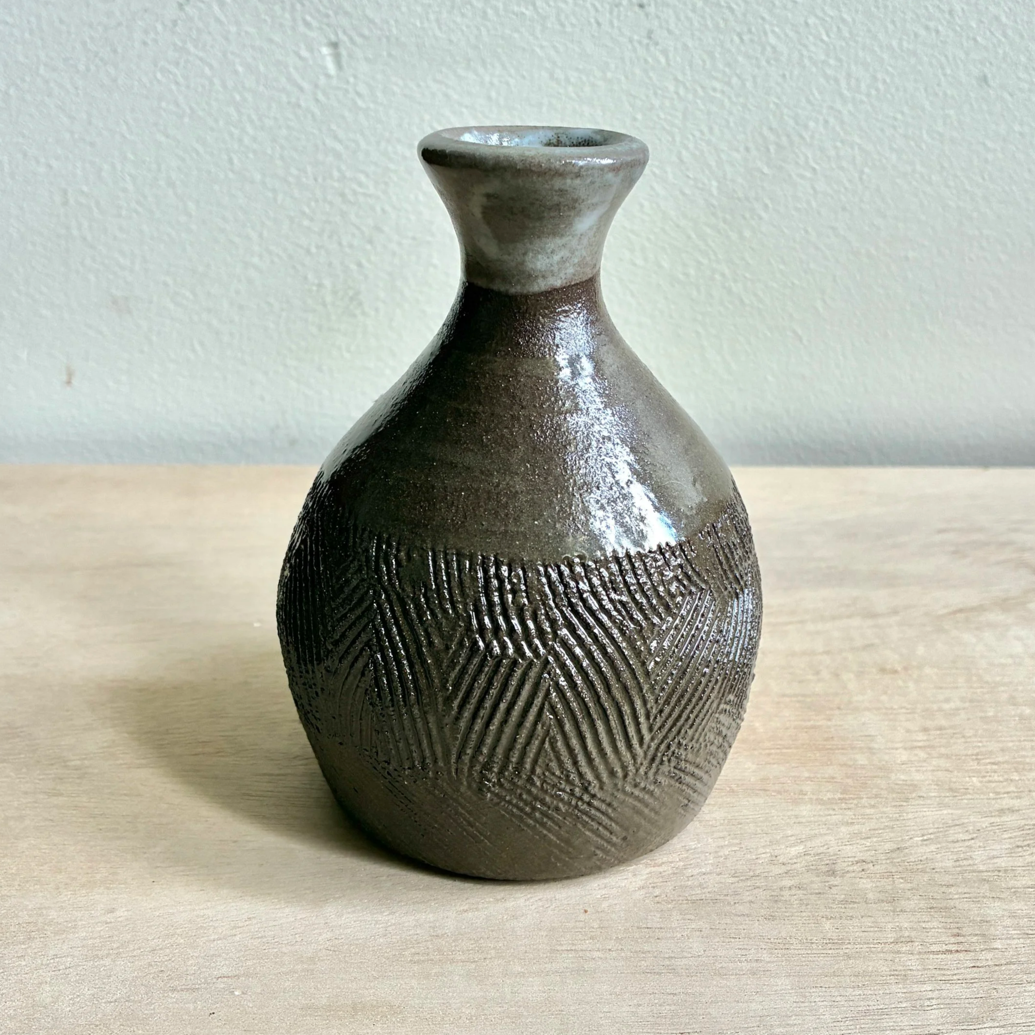 Close-up of a ceramic vase with a textured black pattern on the lower half, a smooth brown middle section, and a glossy, slightly flared neck against a plain wall background.