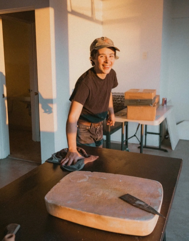 Young person with a cap smiling and cleaning a large wooden cutting board on a table in a room with boxes and a chair in the background.