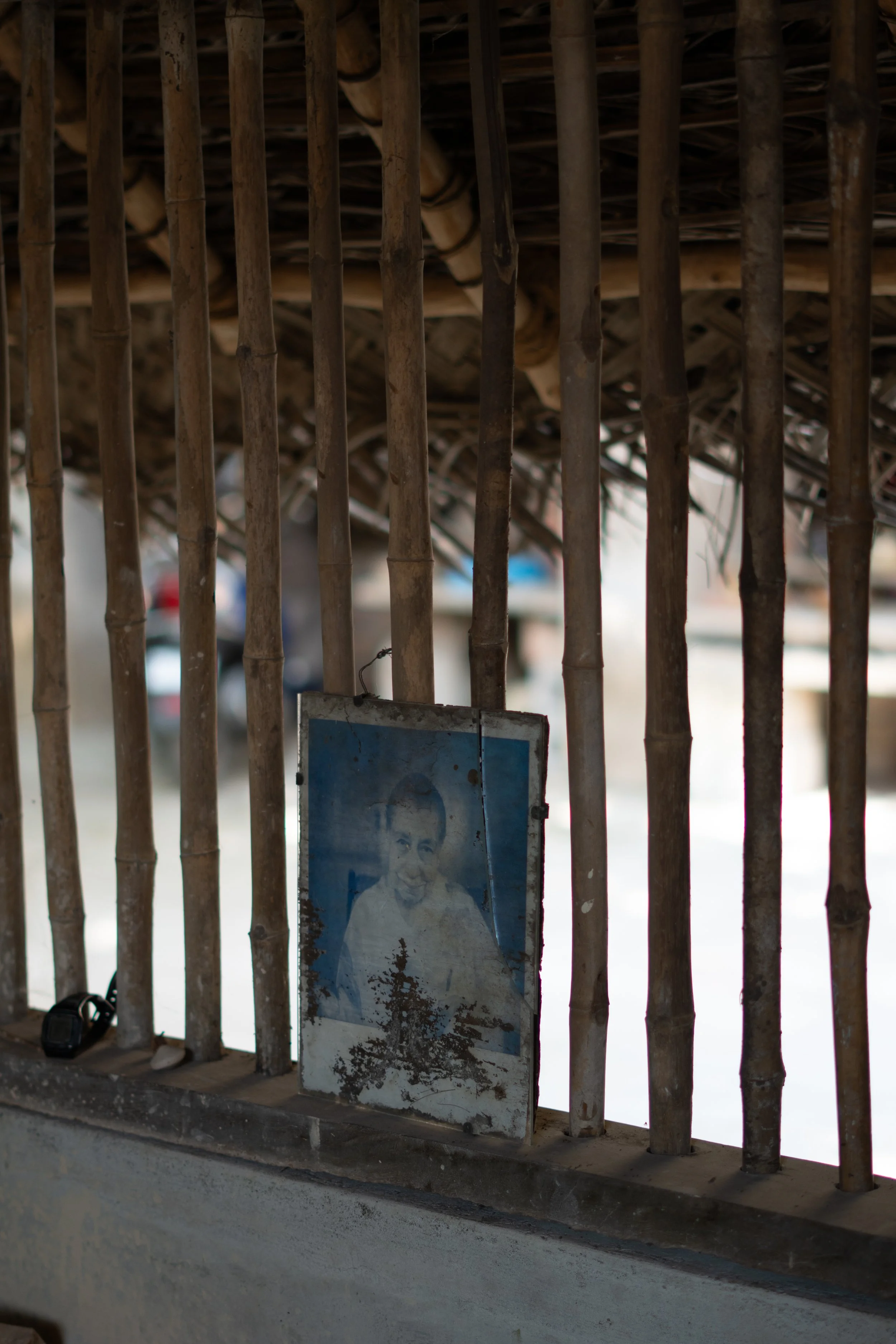 A weathered poster of an elderly man with gray hair, wearing a white shirt, hangs on a bamboo fence. The background is blurred, with some objects visible and the fence is made of vertical bamboo sticks with a horizontal support.