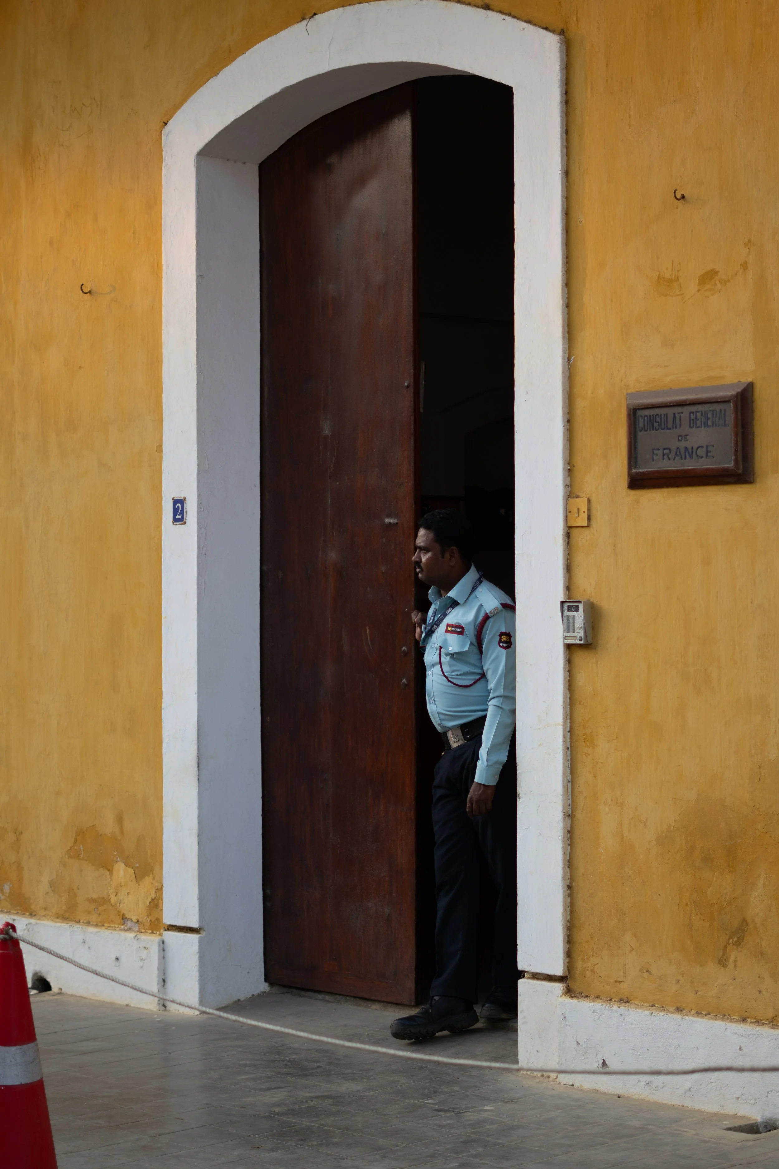 A man in a security uniform standing in the doorway of the French Consulate, with a yellow wall and a sign that says 'Consulat General de France'