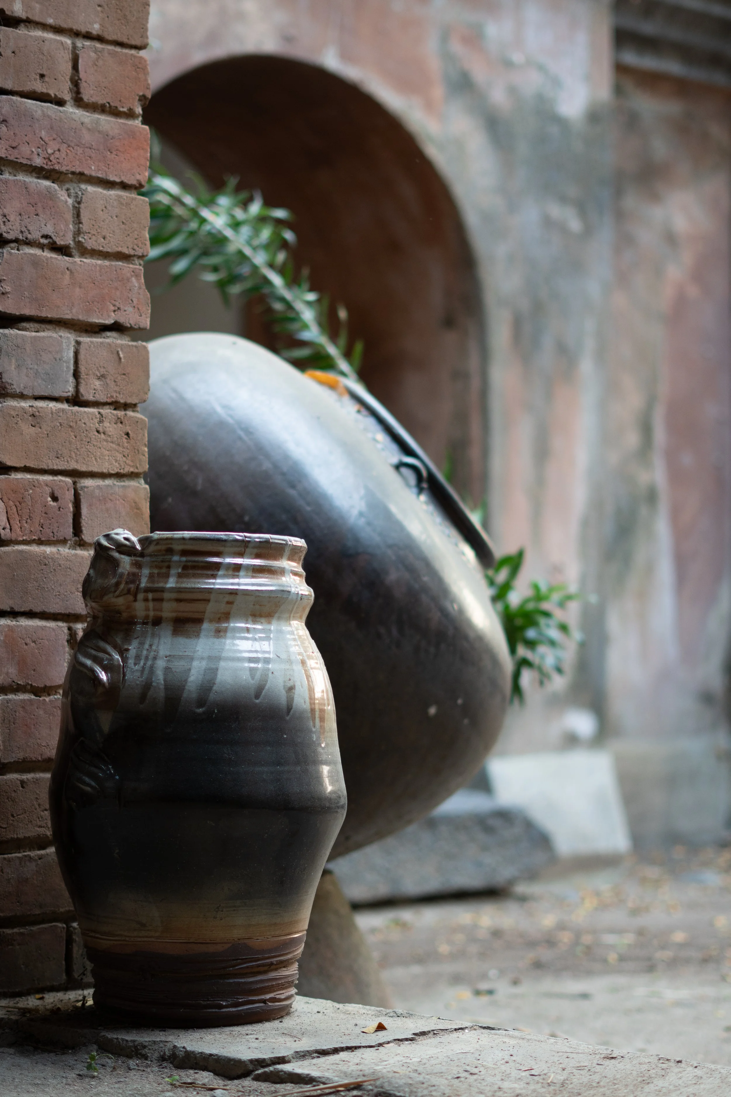 A ceramic vase with a striped pattern in shades of brown and white placed on the ground outside, near a brick wall. In the background, there are large metallic pots with plants and a brick archway, with a rustic outdoor setting.