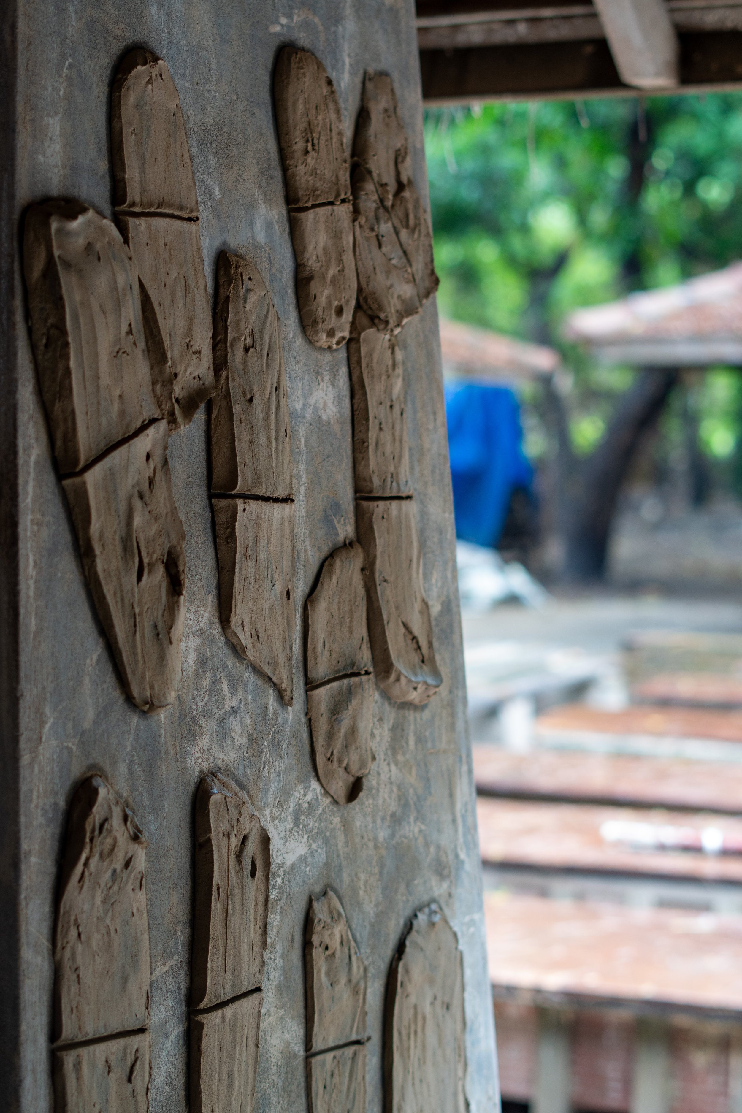 Several clay molds of shoes drying on a concrete wall, outdoors with blurred green trees and wooden structure in the background.