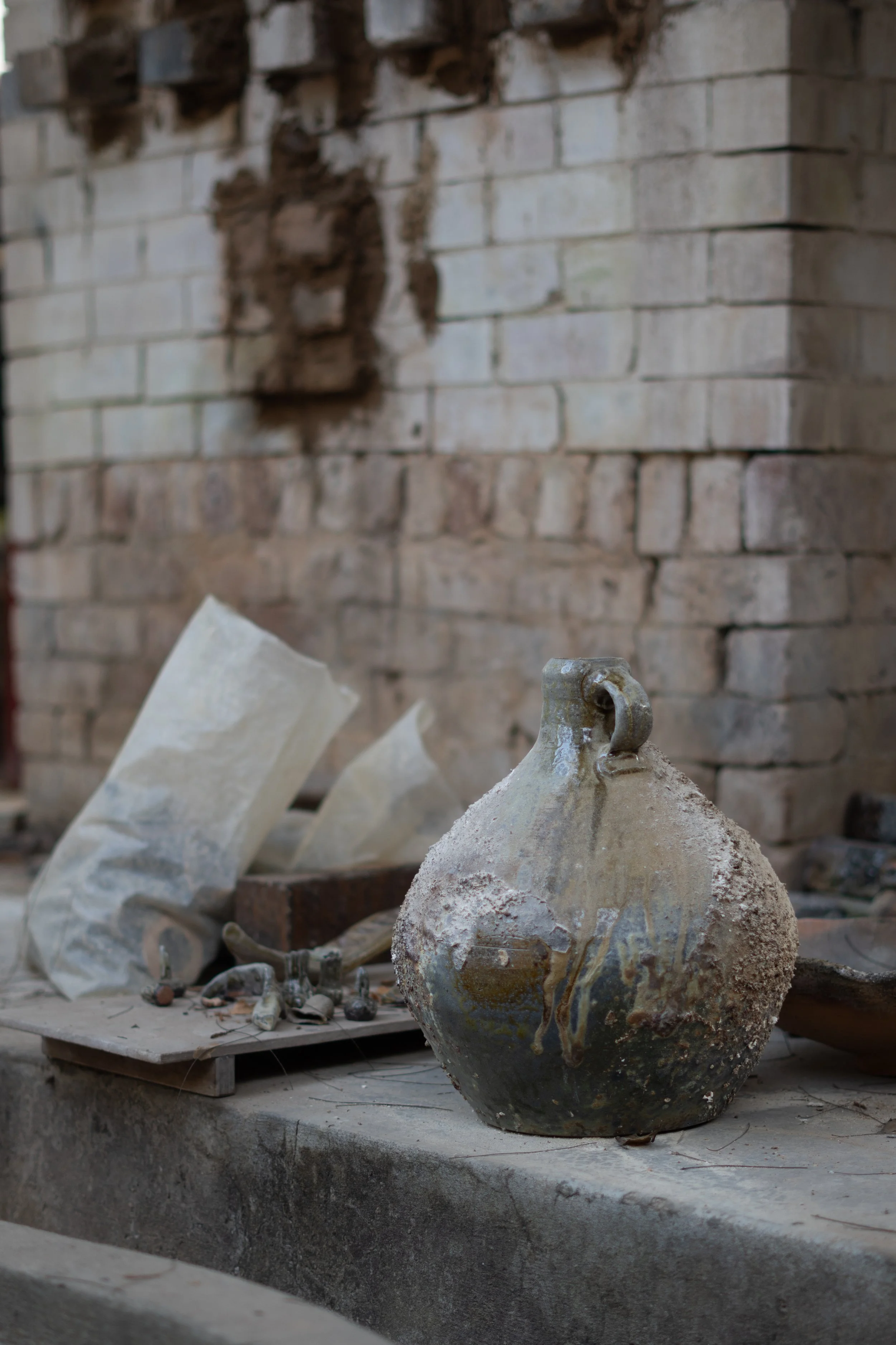 An old, weathered ceramic vessel or pot with a handle, sitting in front of a brick wall. In the background, there are other ceramic pieces and tools, indicating a pottery workshop or studio.