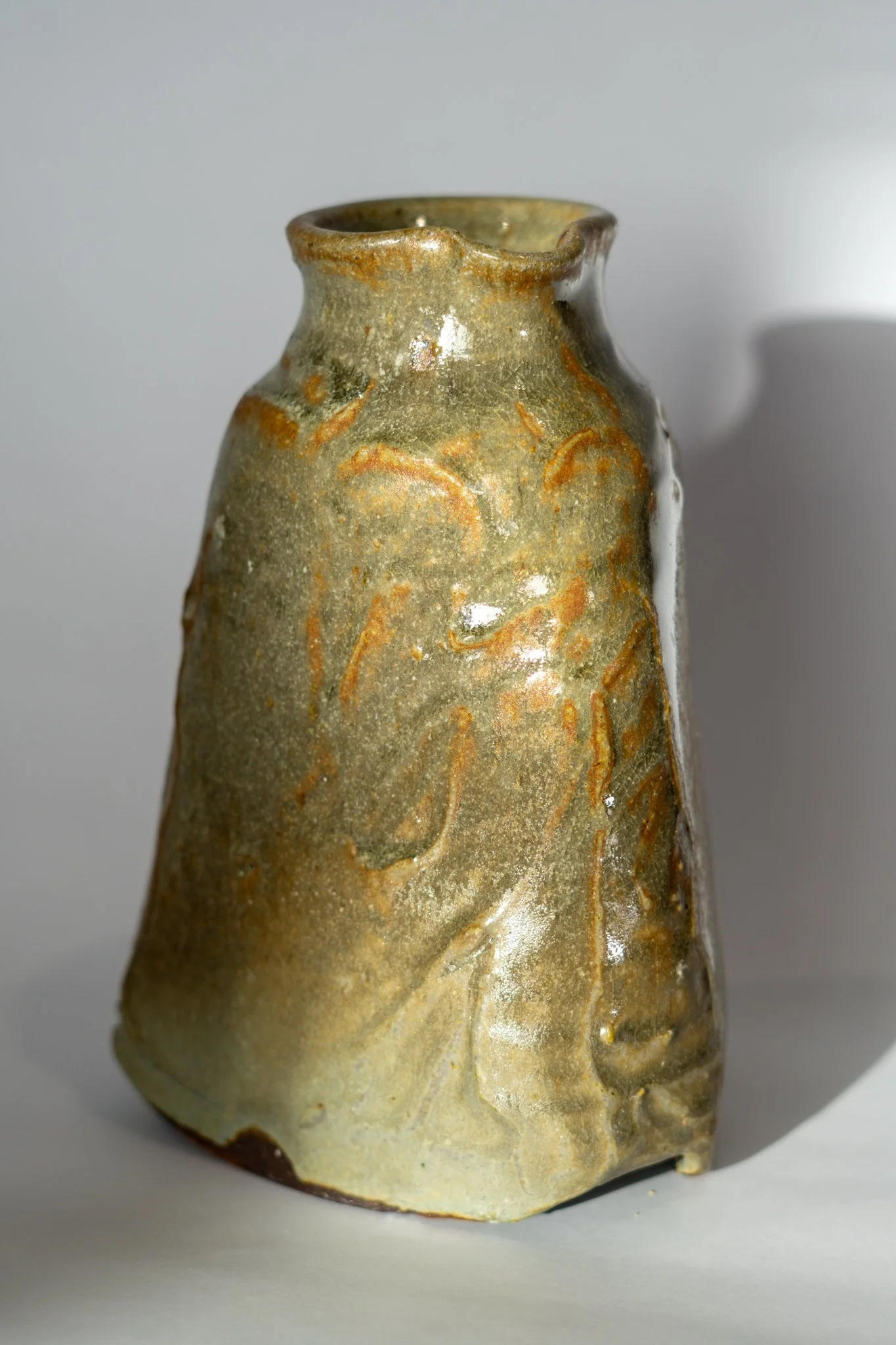 Close-up of a small, ceramic pottery vessel with a slightly flared, uneven rim, glazed in earthy tones with a textured surface and subtle orange-brown streaks, set against a plain gray background.