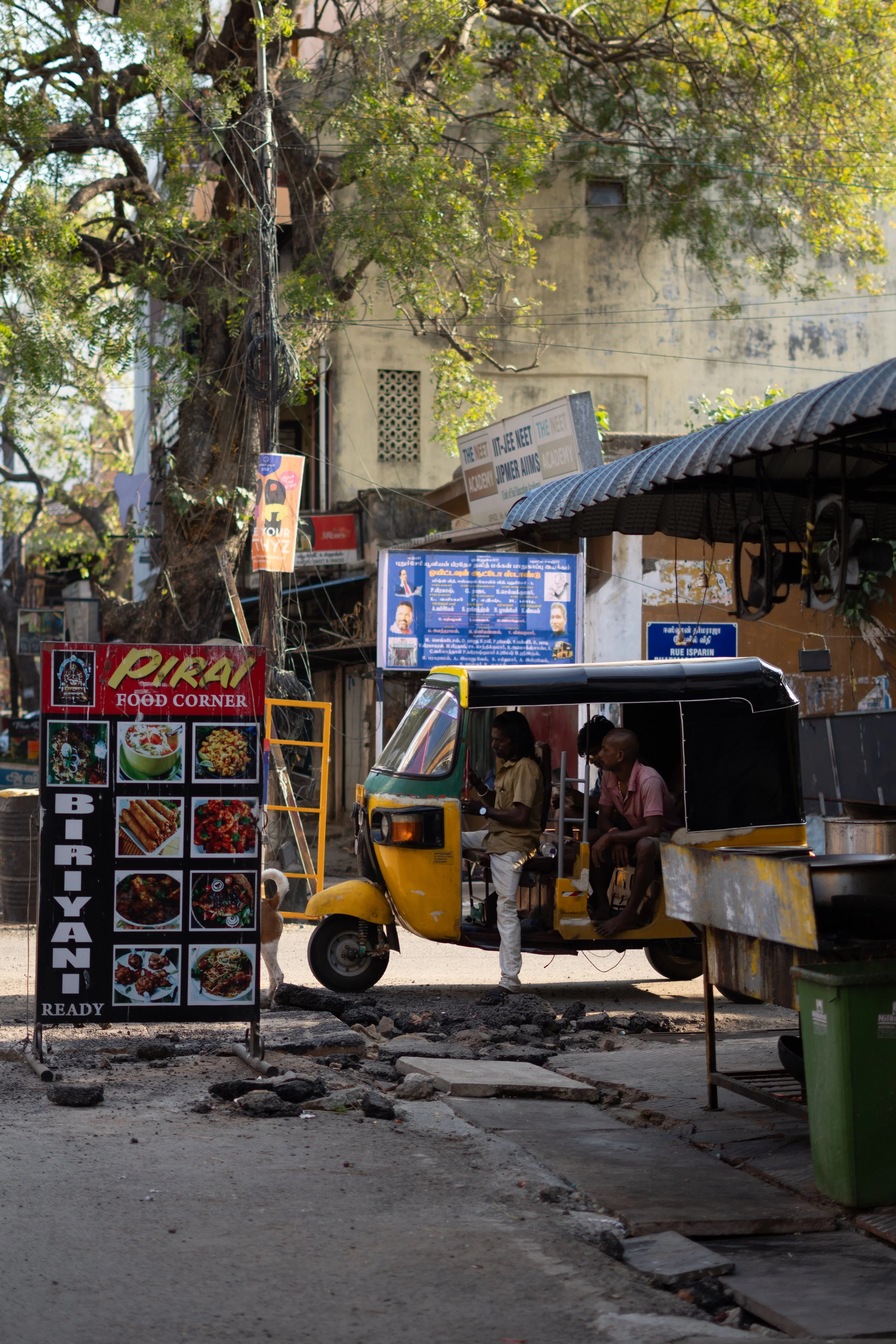Street scene with a yellow auto rickshaw, a man standing beside it, and a signboard advertising Indian food. There are trees, building walls, and overhead wires in the background, with some uneven pavement and debris on the ground.