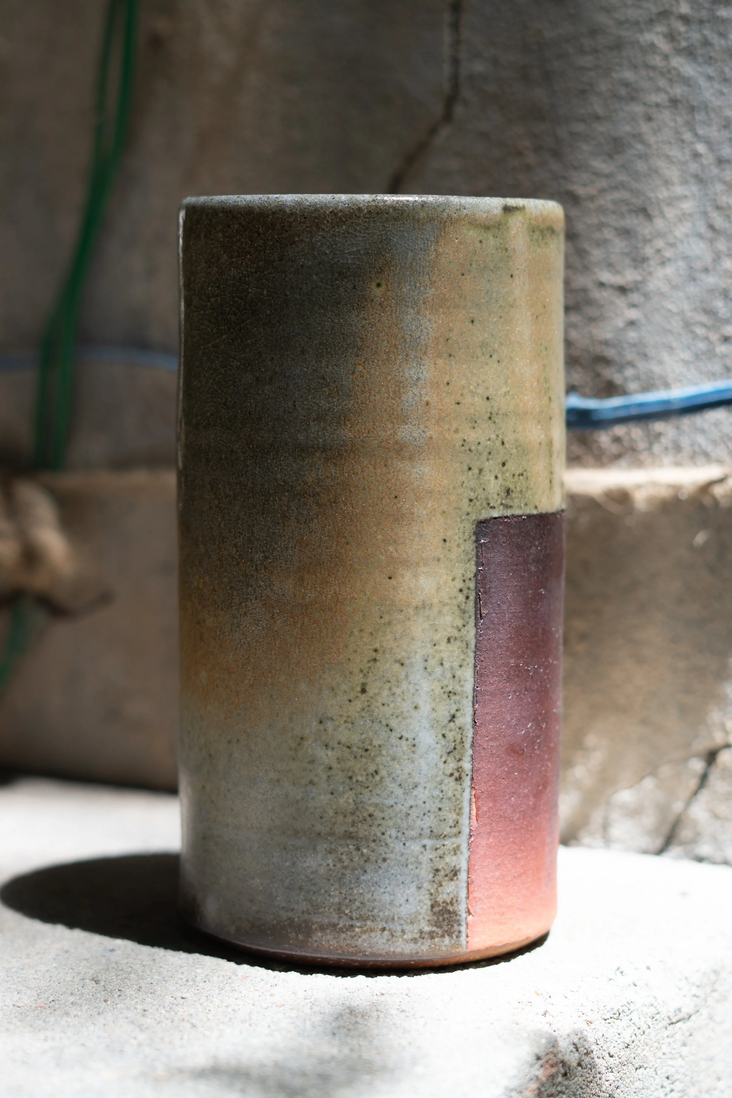 Close-up of a ceramic or concrete cylindrical object resting on a sandy surface, with a grassy and stone-textured background.