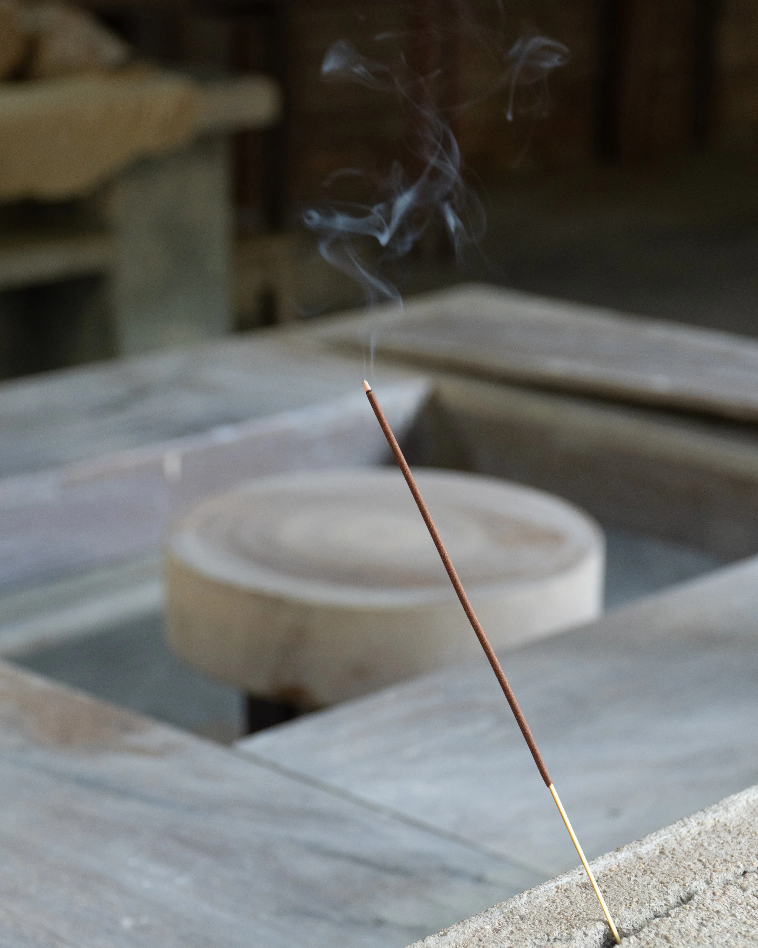 Incense stick burning on a surface with smoke curling upward, background showing blurred chairs and table.
