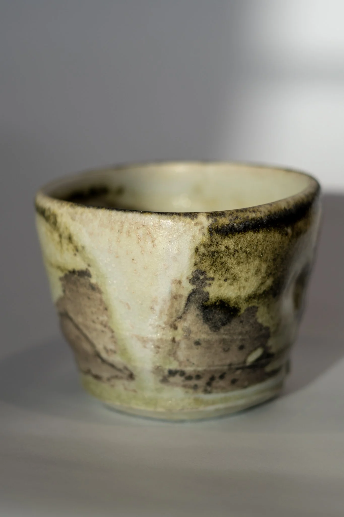 Close-up of a ceramic cup with a beige glaze and dark irregular spots, set against a neutral background.