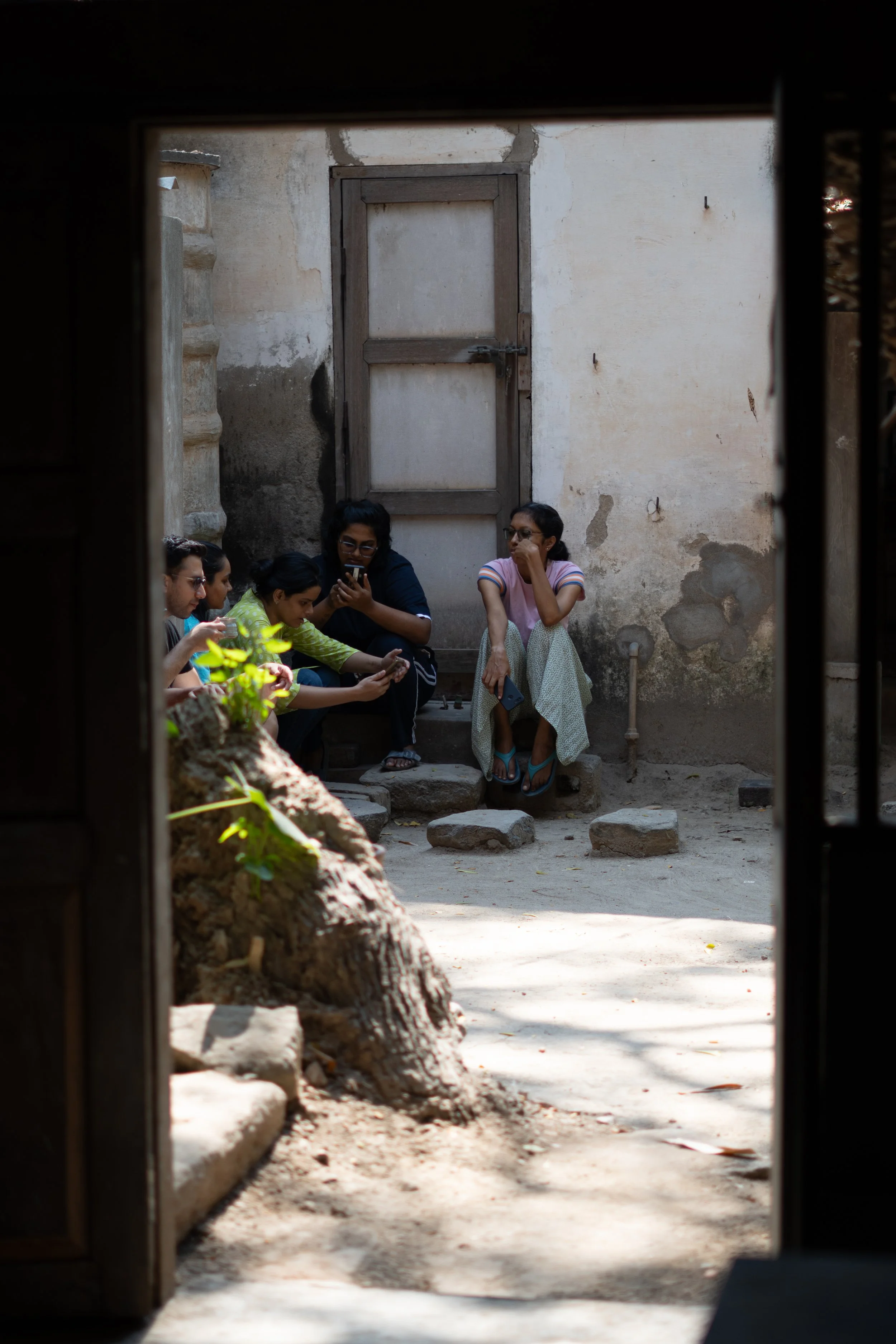 A group of five people sitting outside in a shaded courtyard, looking at their phones, with a woman in a pink shirt and long skirt sitting on a stone step, and a large tree trunk in the foreground.