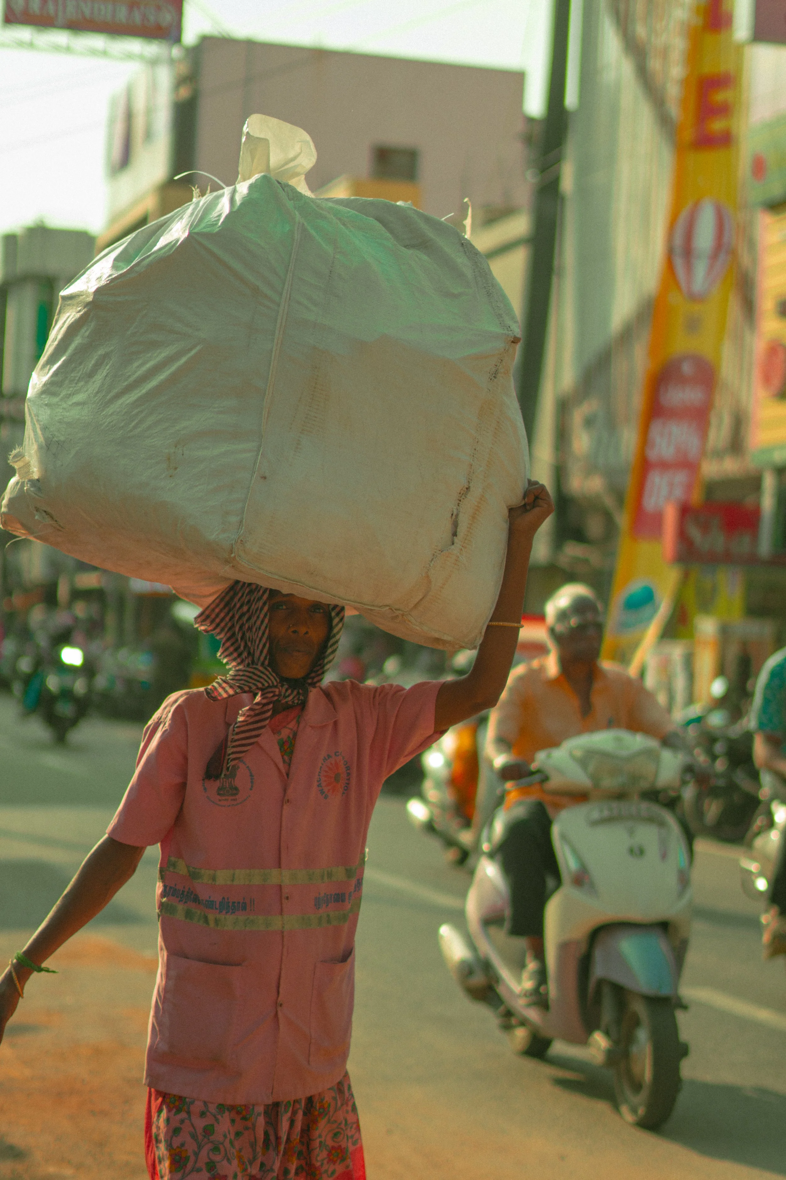 A woman wearing a pink shirt, patterned leggings, and a head covering carries a large bag on her head while walking on a busy street with motorcycles and colorful signs in the background.